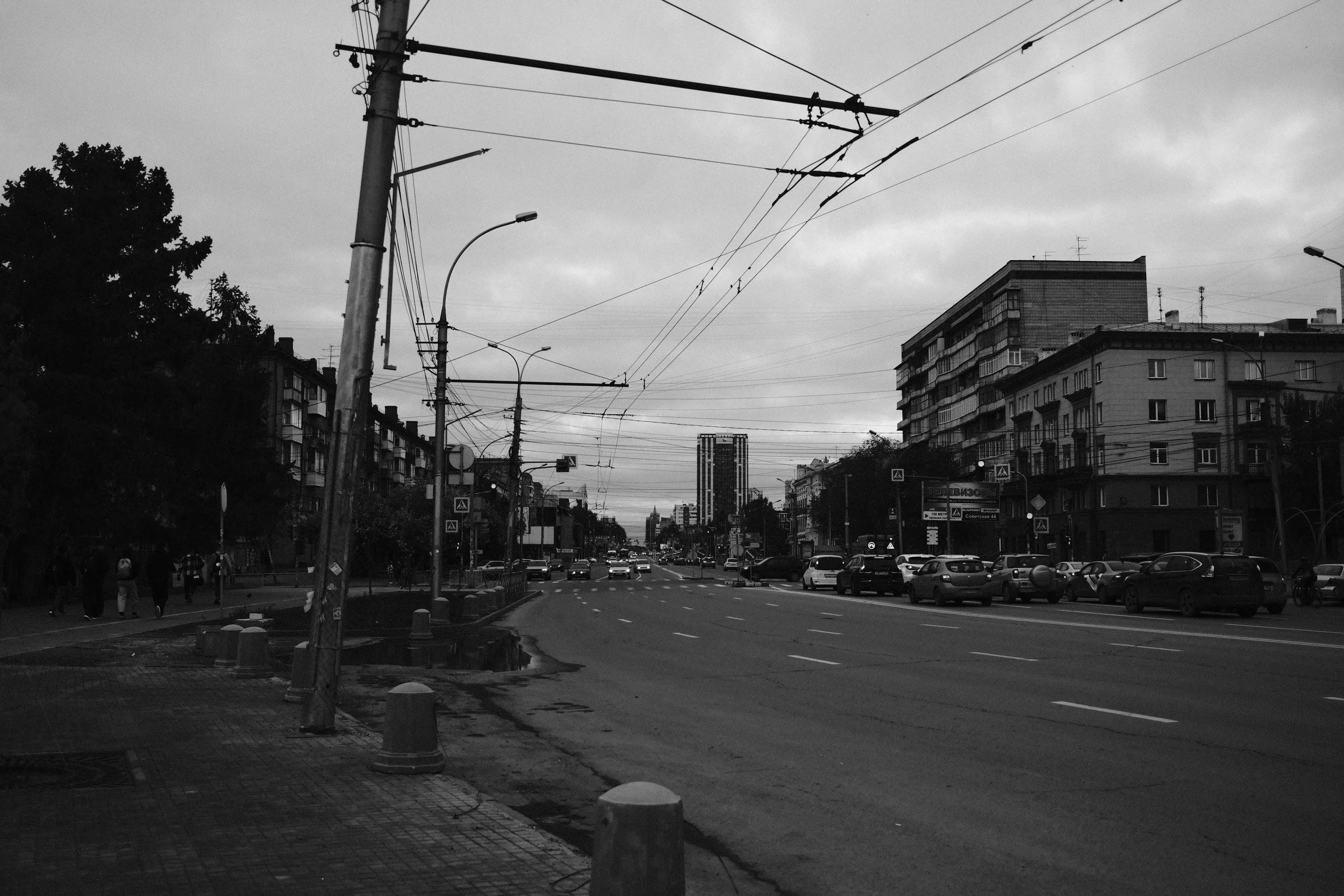 A city street scene in black and white showing cars parked along the right side of the street, with street and traffic lights, overcast sky, and buildings on both sides, including a tall building in the distance.