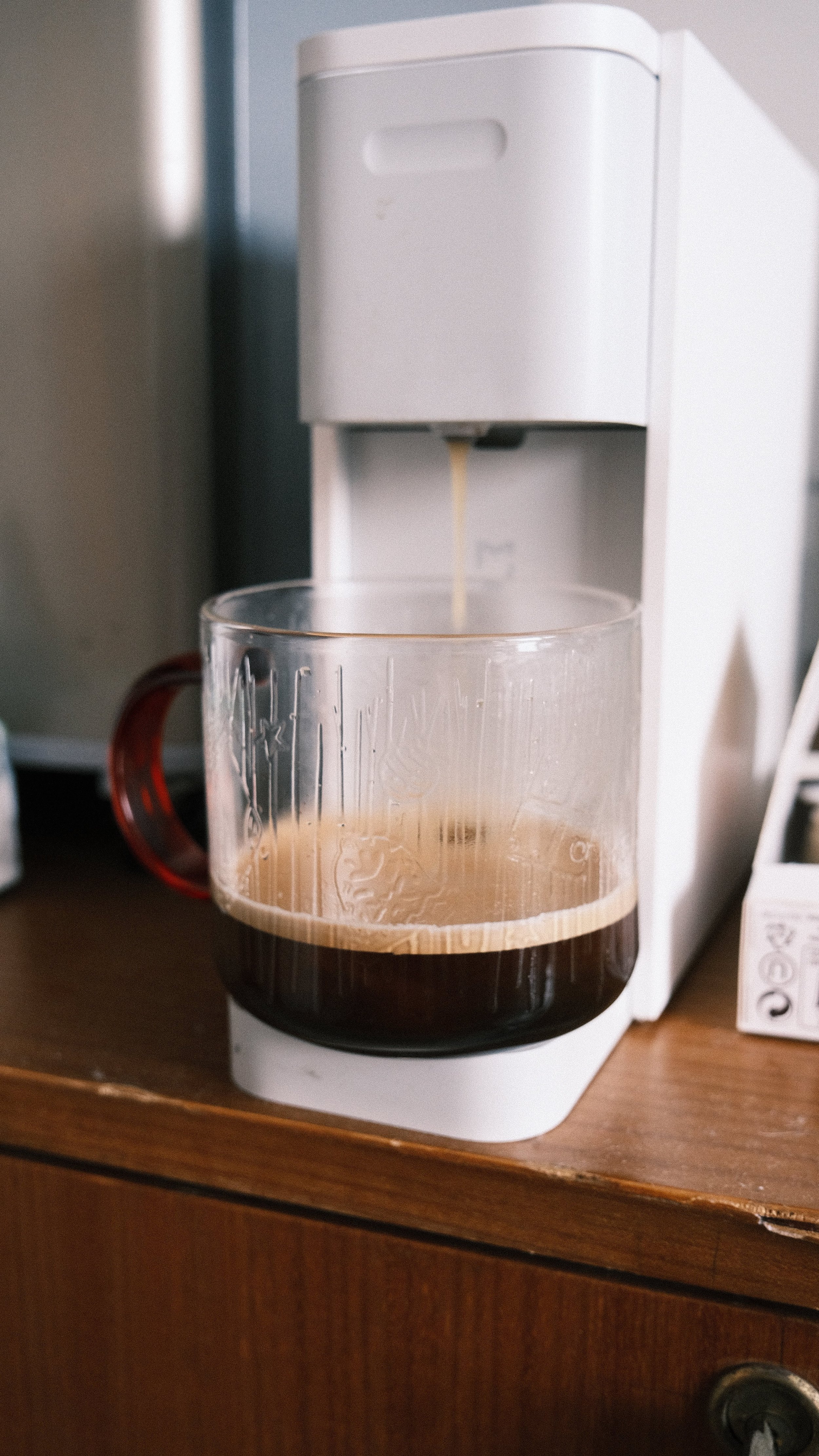 Close-up of a white coffee maker on a wooden surface, brewing coffee into a clear glass mug with a red handle.