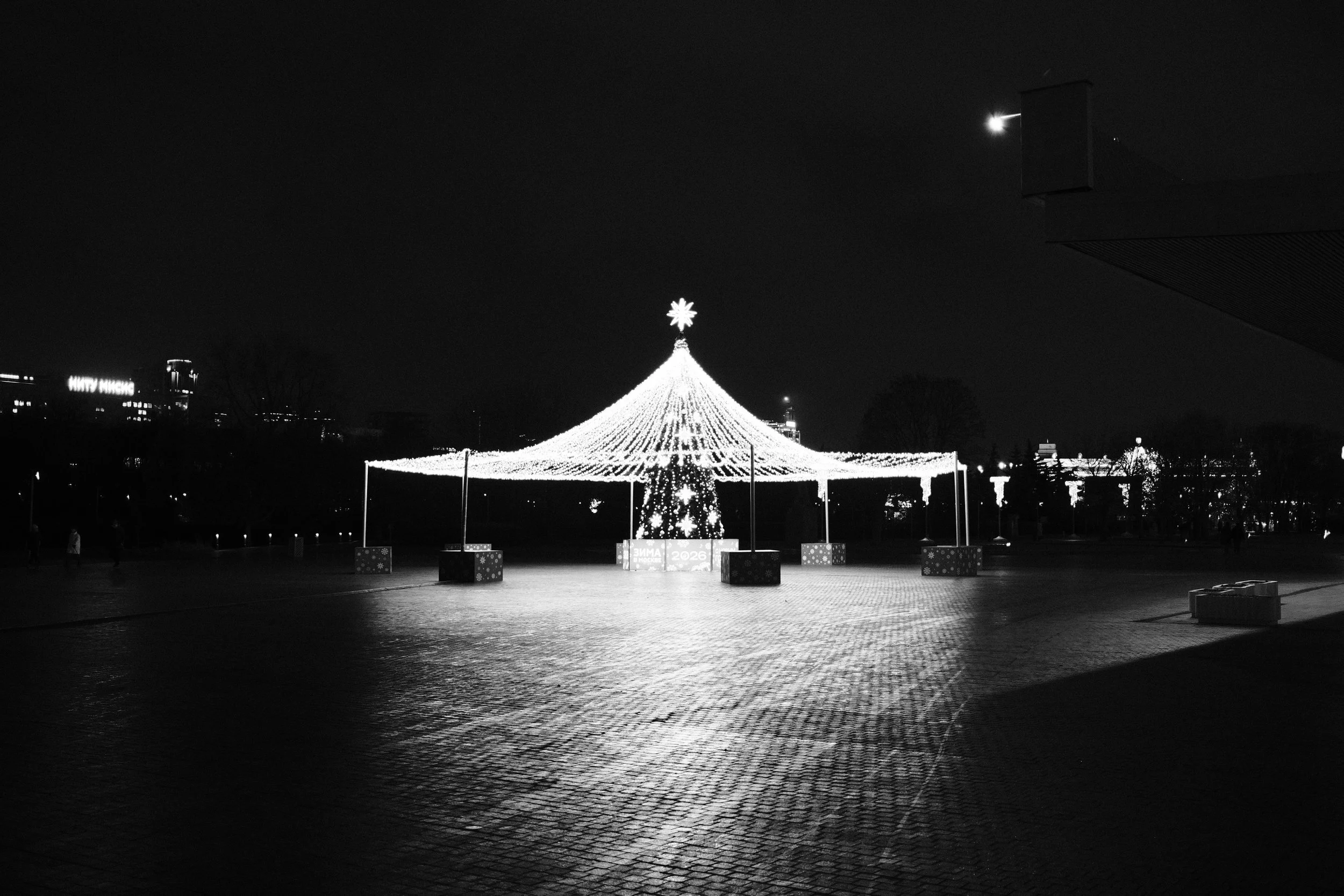 Black and white photo of a large, illuminated Christmas tree decoration with strings of lights, shaped like a cone, topped with a star, outdoors at night.
