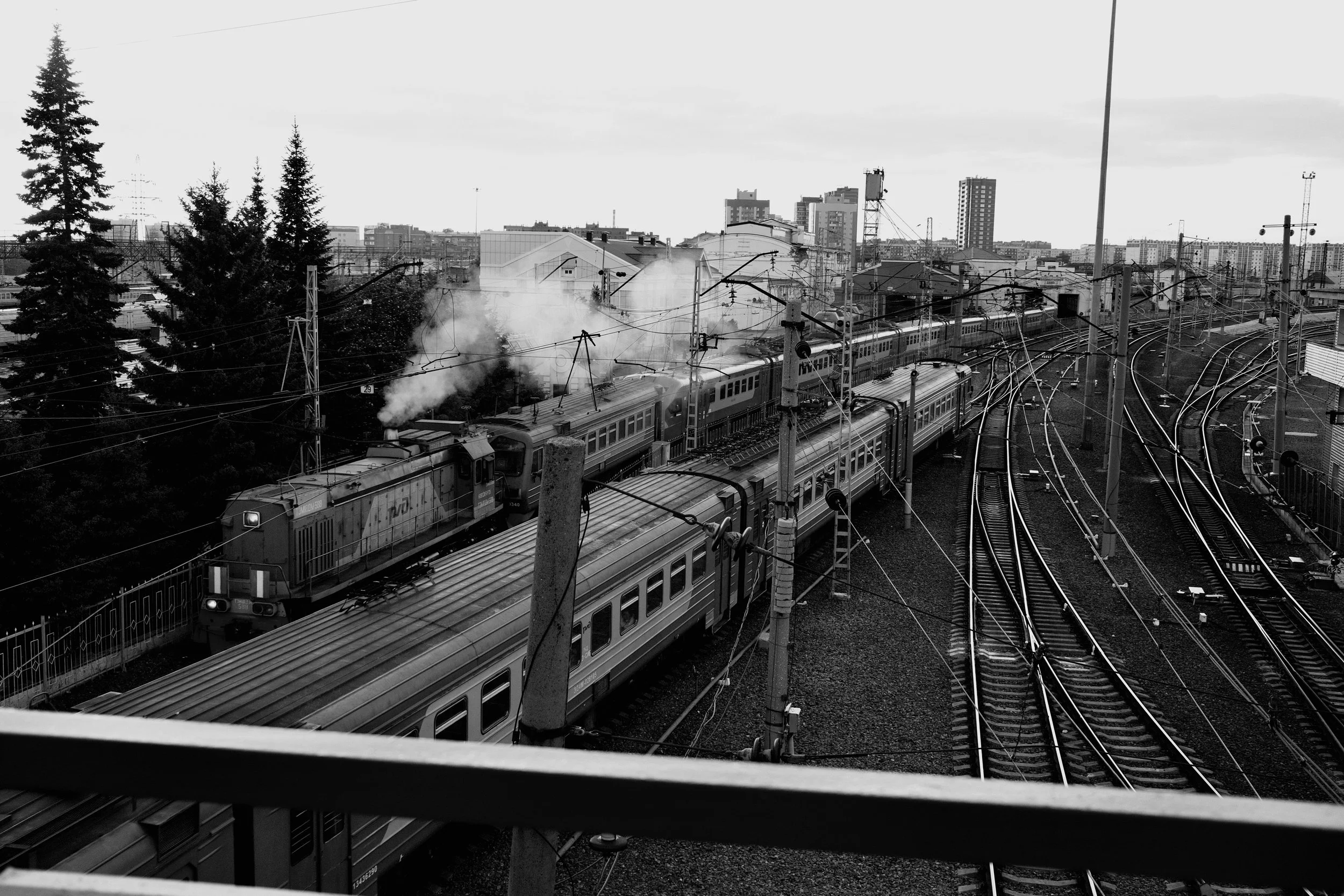 Black and white photo of a train on tracks, with smoke emitting from the engine, set in an urban area with city buildings in the background.