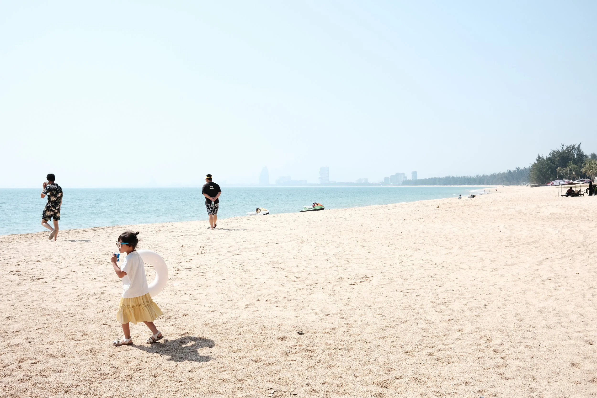 People enjoying a sunny day at the beach, with children playing and adults relaxing near the shoreline, boats on the water, and distant buildings visible.