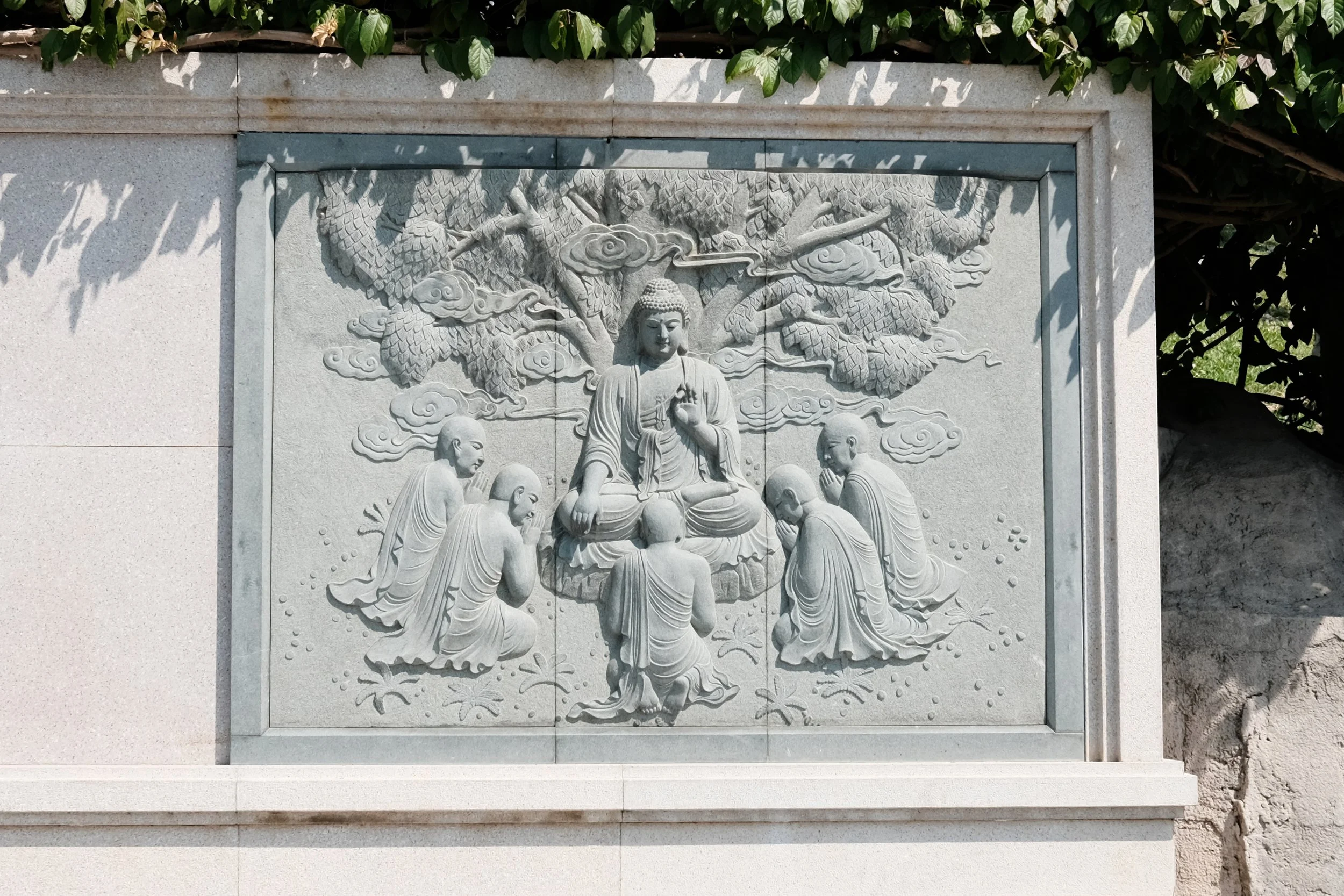 A carved stone relief depicting a seated Buddha surrounded by six monks or disciples, under a large tree with detailed leaves and clouds in the background.