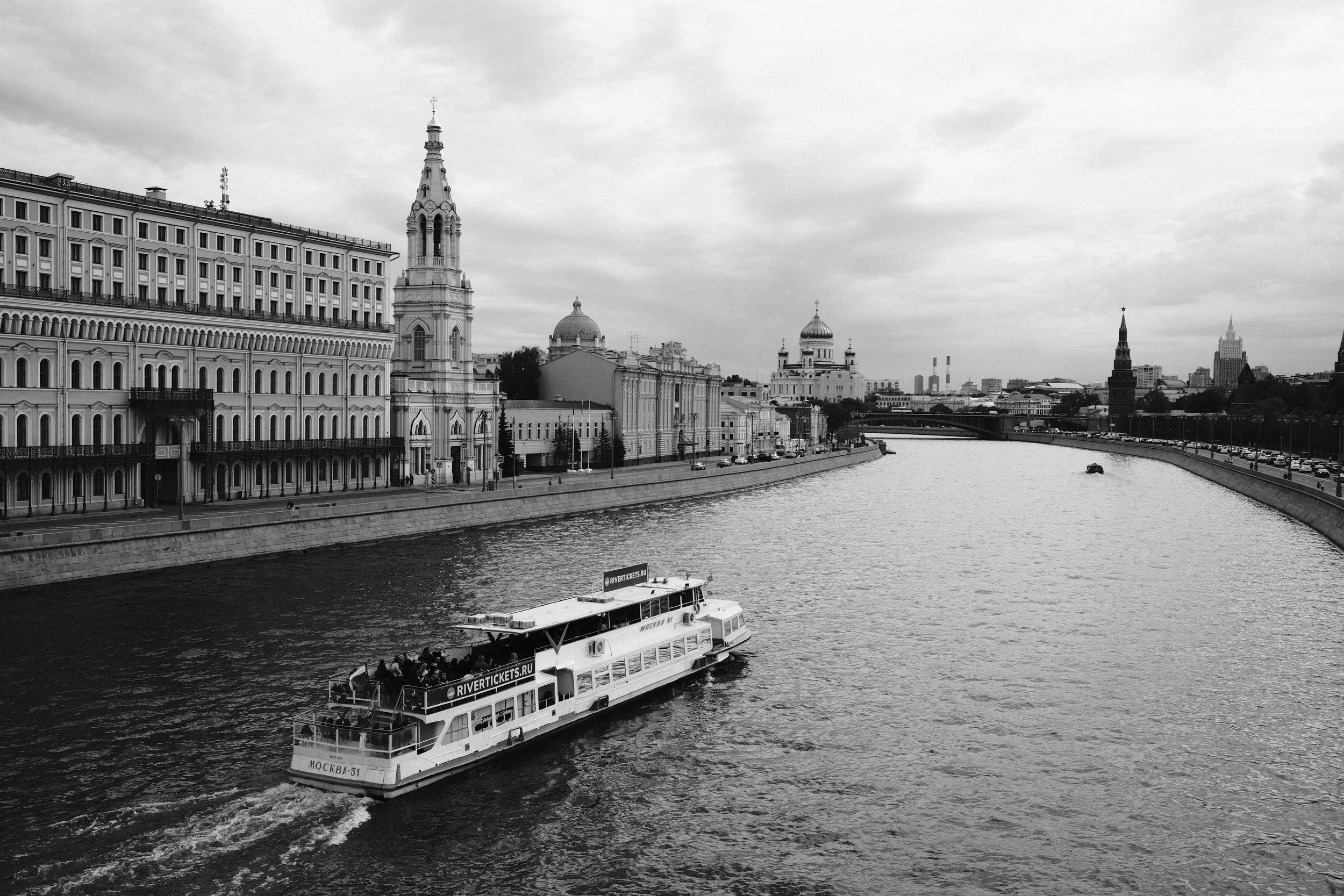 A boat sailing on a river in a city with historic buildings and church domes under a cloudy sky.