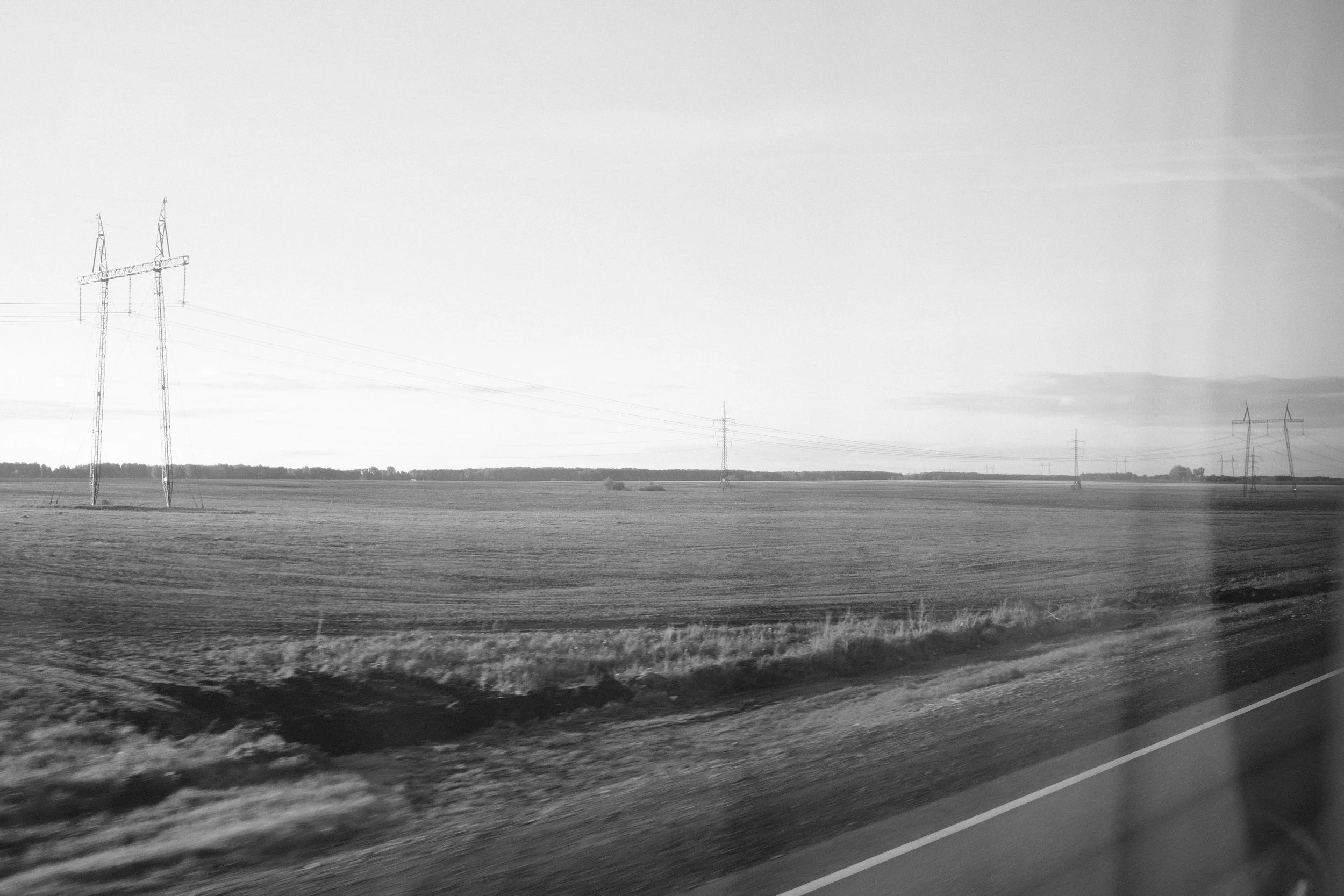 Black and white photo of flat farmland with power lines and transmission towers, taken from a vehicle on the highway.
