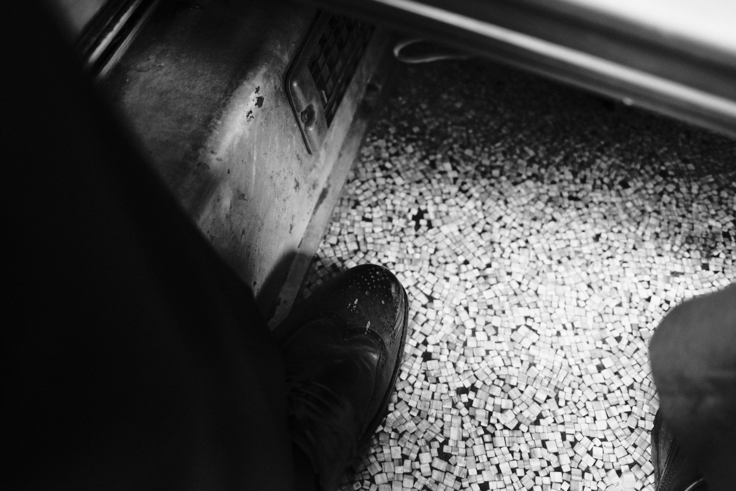 A person's foot in a shoe standing on a floor covered with small mosaic tiles, with part of a metal panel and a ventilation grill visible at the top left of the image.