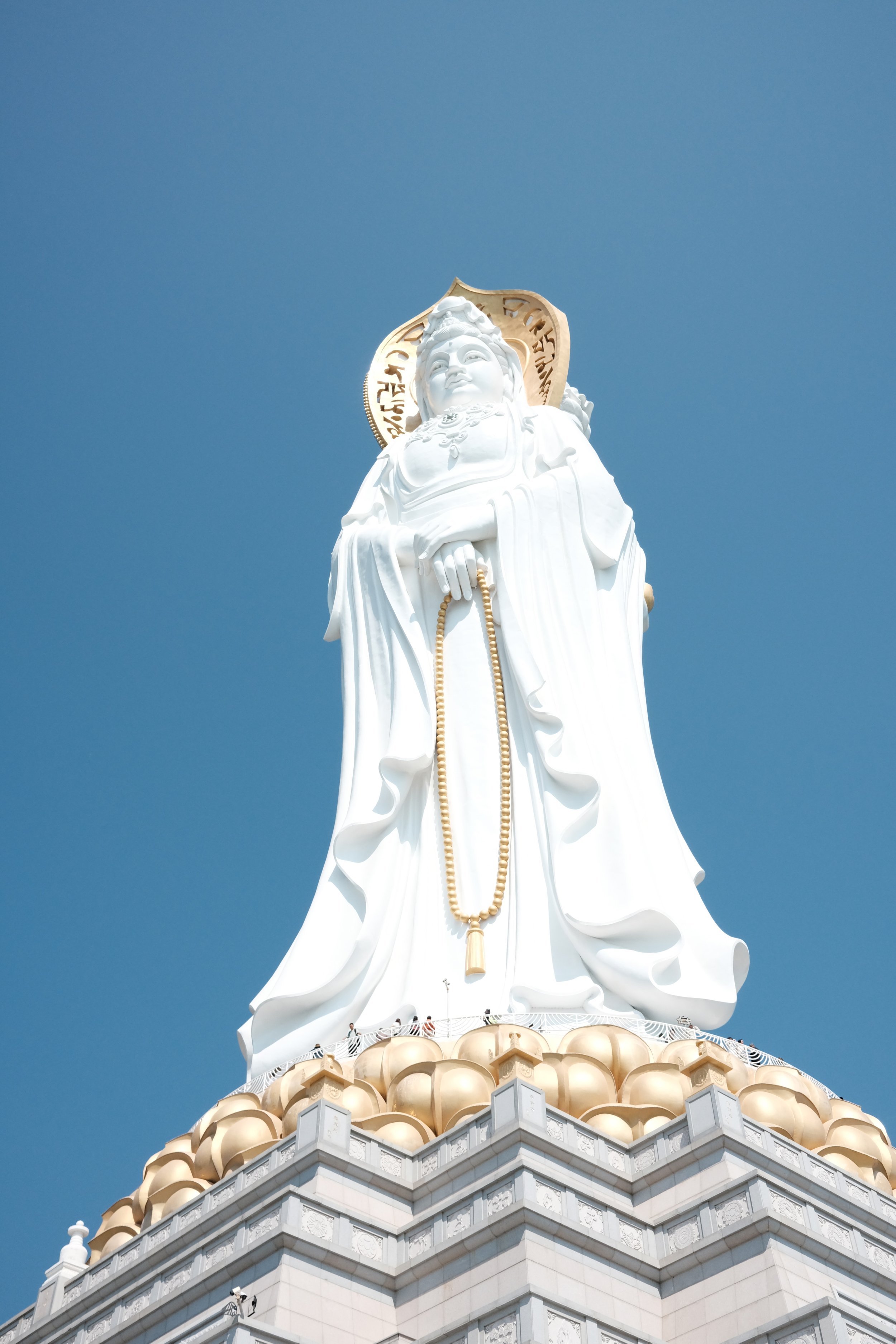 A tall white statue of a religious figure standing on a decorated base with a blue sky in the background.