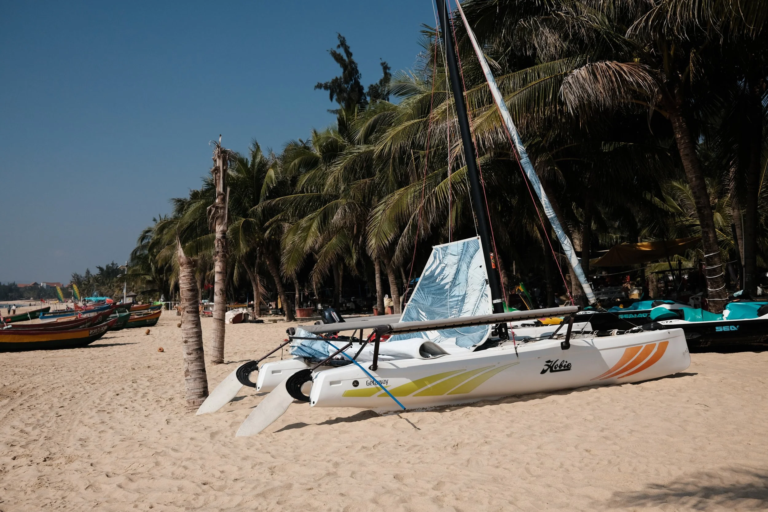 A sailboat resting on sandy beach near palm trees with other boats in the background.