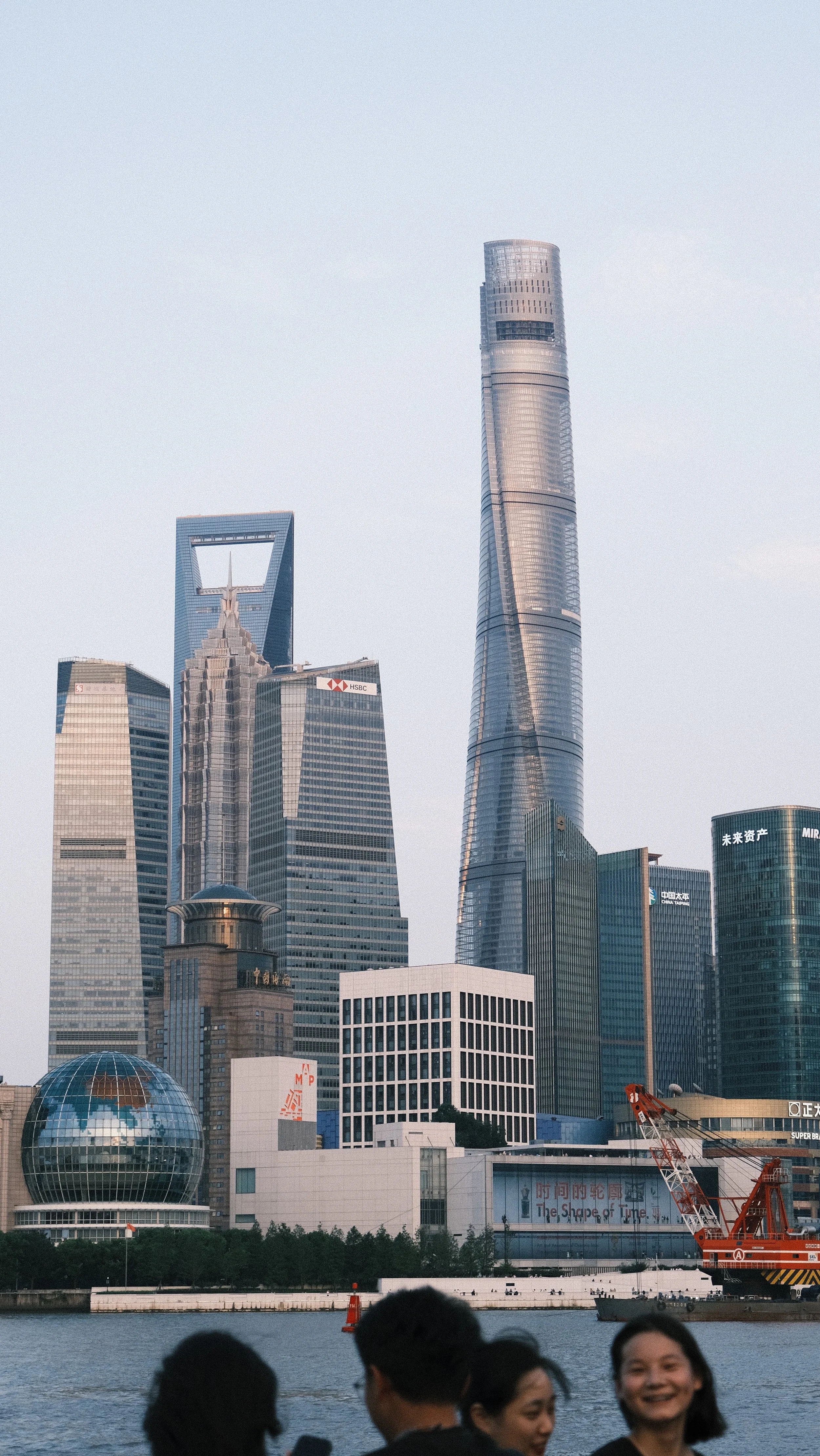 View of the Shanghai skyline with tall skyscrapers, including the Shanghai Tower, the Jin Mao Tower, and the Shanghai World Financial Center, with people in the foreground near a river.