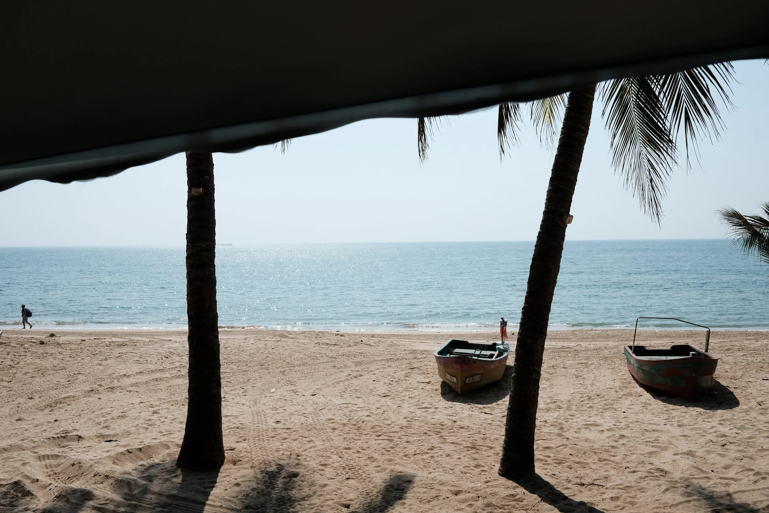 View of a sandy beach with two small boats resting on the sand, two tall palm trees between the boats, and a person walking along the shoreline in the distance, with calm ocean water and a clear sky in the background.
