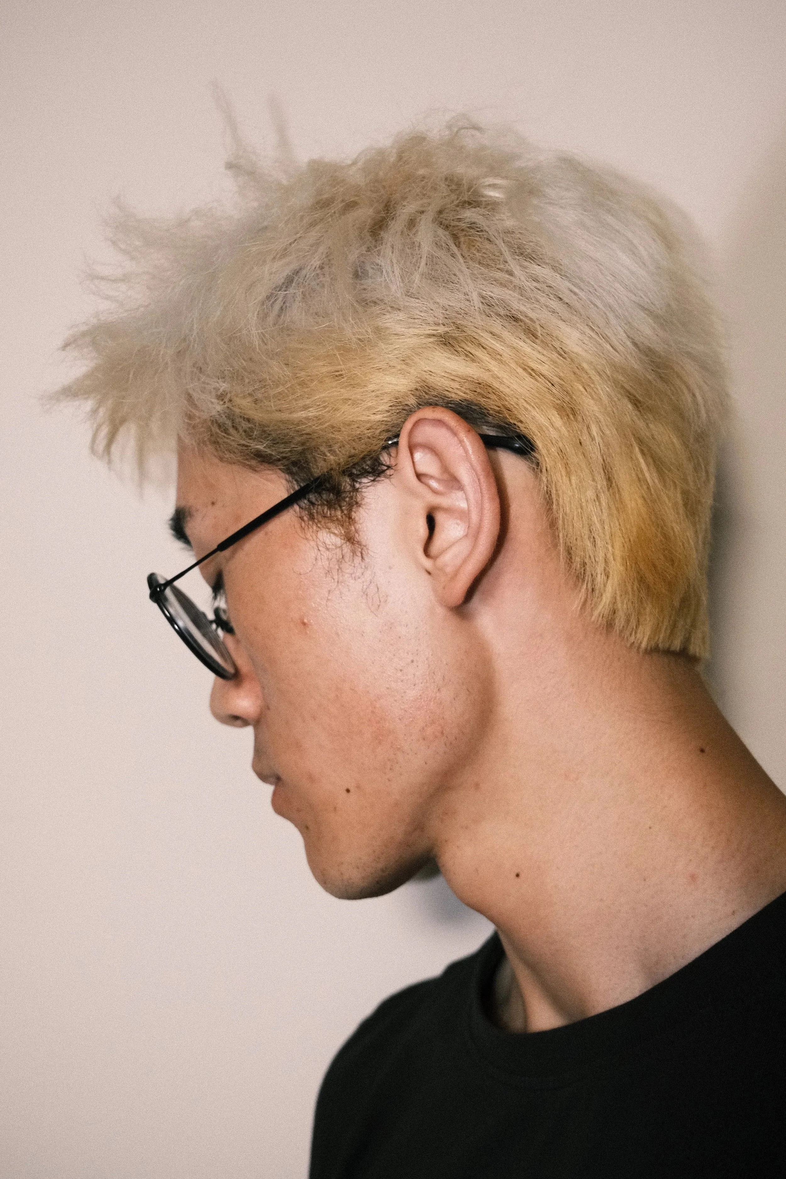 Close-up side profile of a young person with bleached blonde spiky hair, wearing black-rimmed glasses, and a black shirt against a plain background.