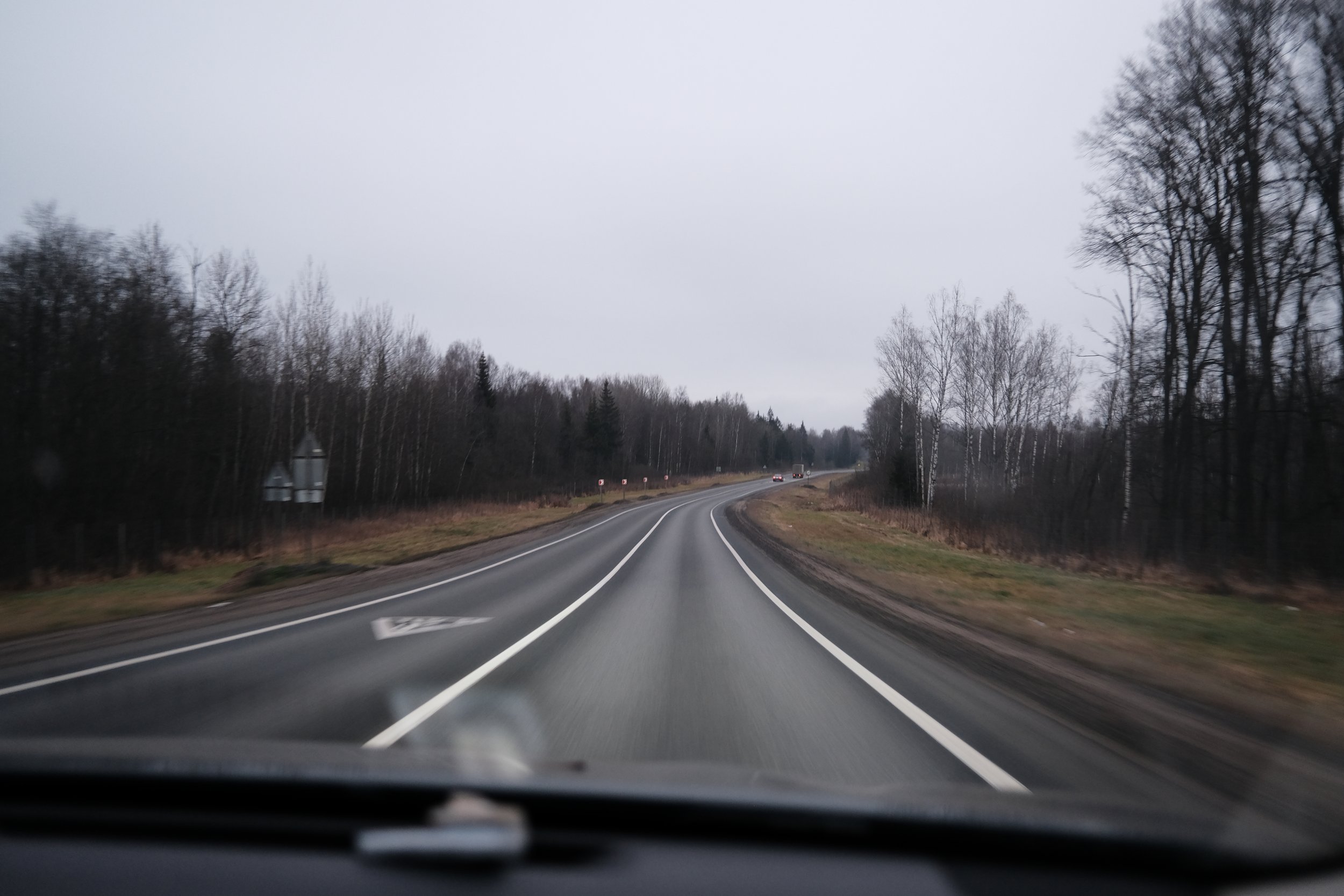 A view of a two-lane road with a double solid line down the middle, curving to the right. The road is bordered by grassy shoulders and leafless trees, with a cloudy, overcast sky overhead.