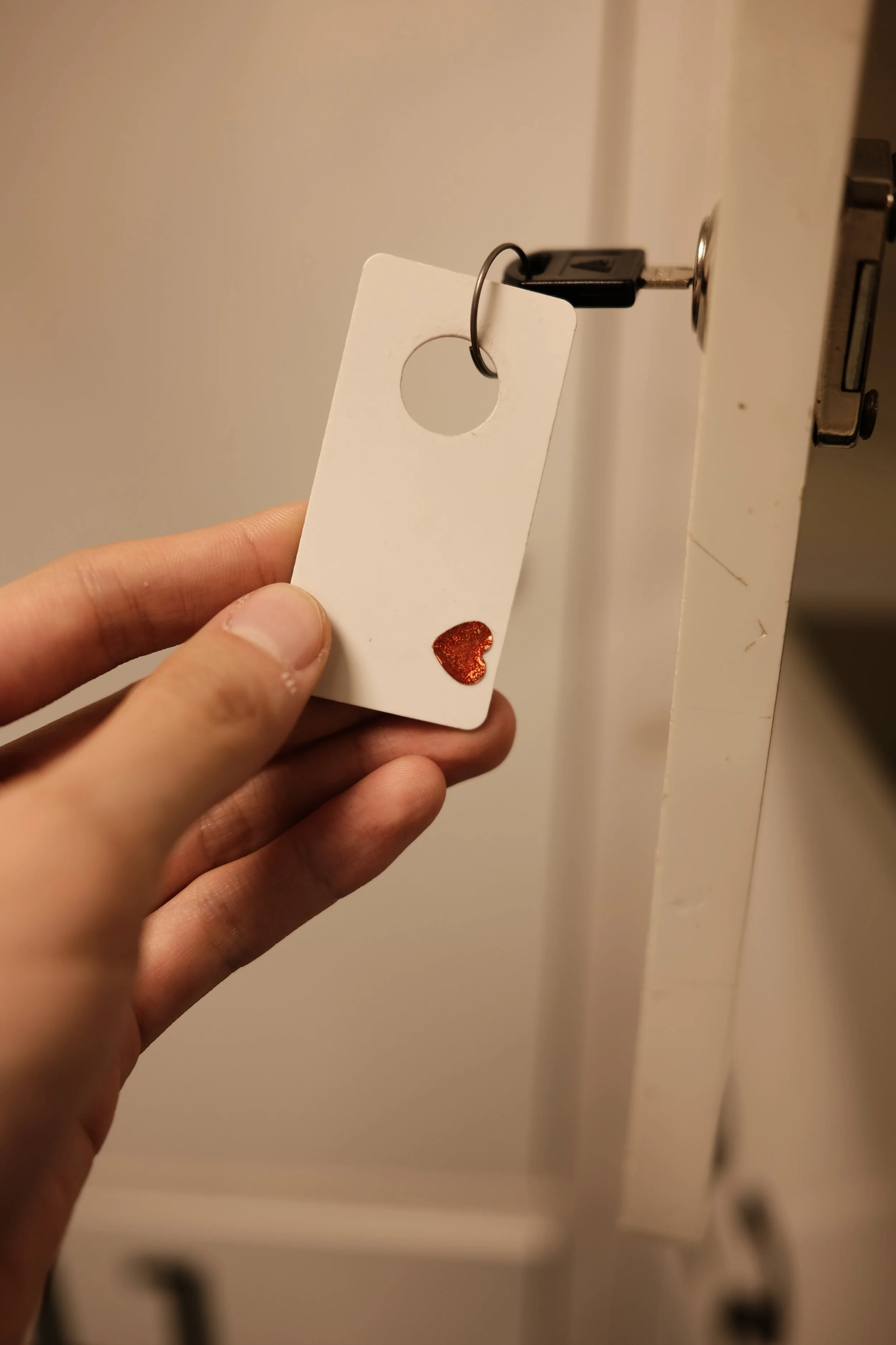 Close-up of a hand holding a white door hanger with a red glitter heart sticker, hanging on a doorknob with a key in the lock.