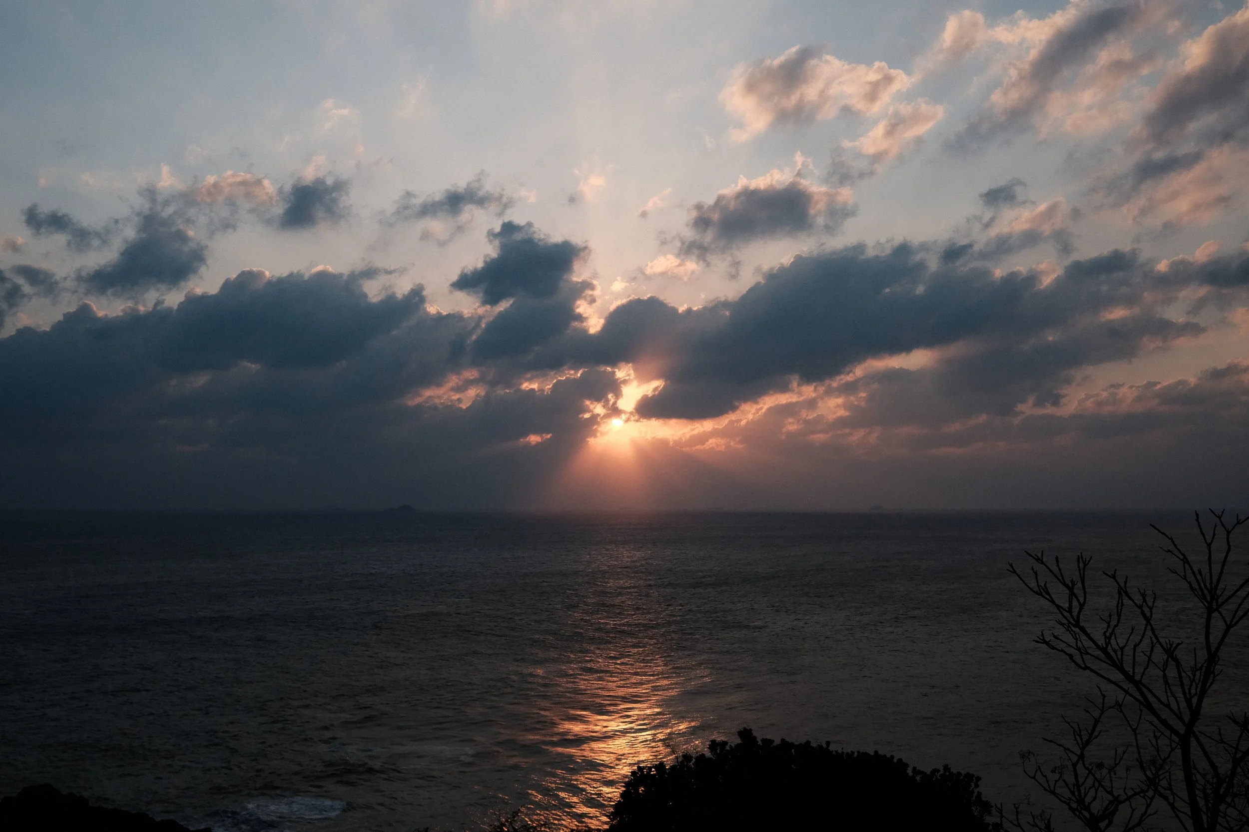 Sunset over the ocean with clouds partially blocking the sunlight and a reflection on the water, with silhouettes of trees and bushes in the foreground.