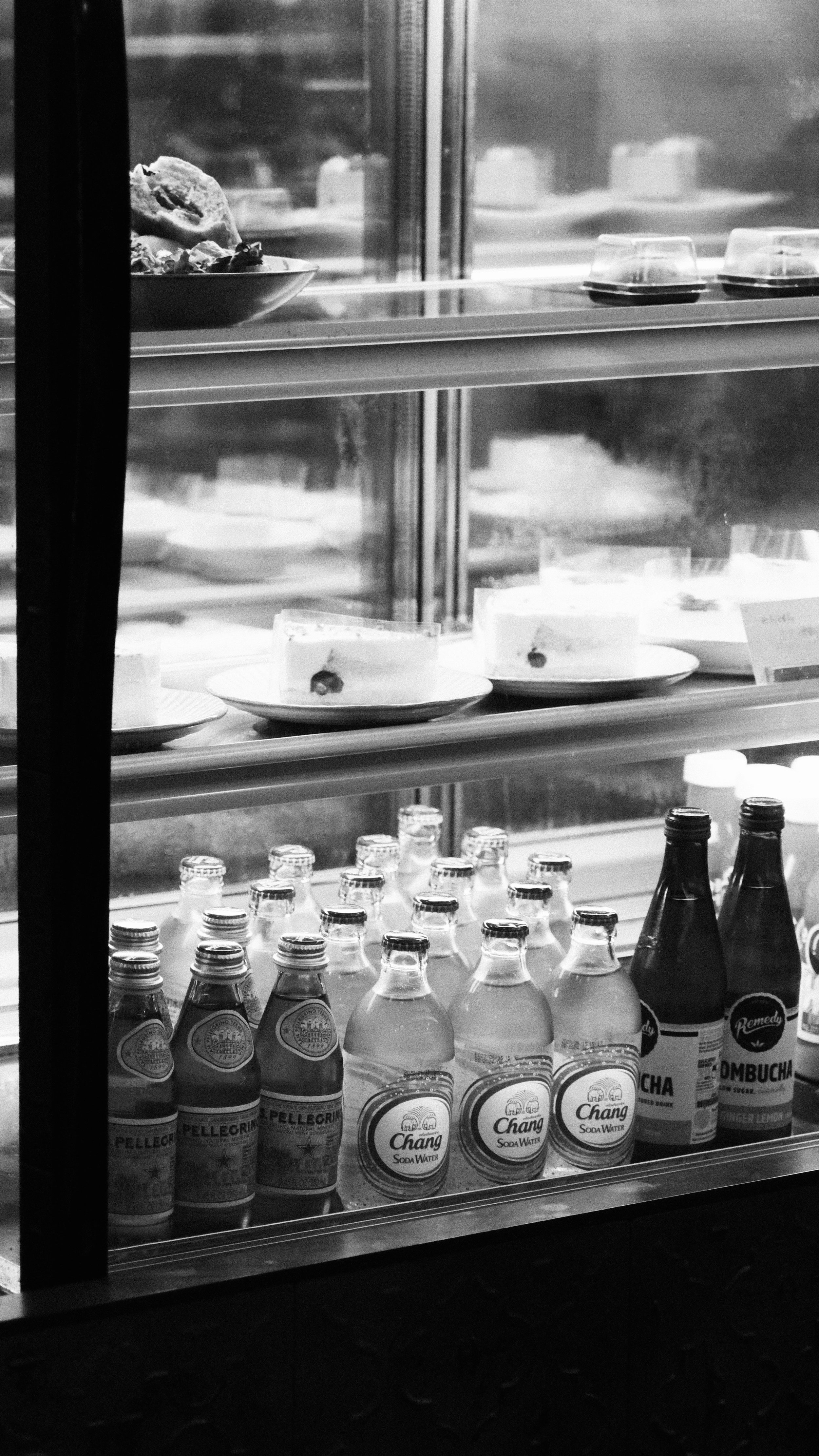 A black and white photo of a refrigerated display case with bottled sodas on the bottom shelf and slices of cake and other desserts on the middle shelf.
