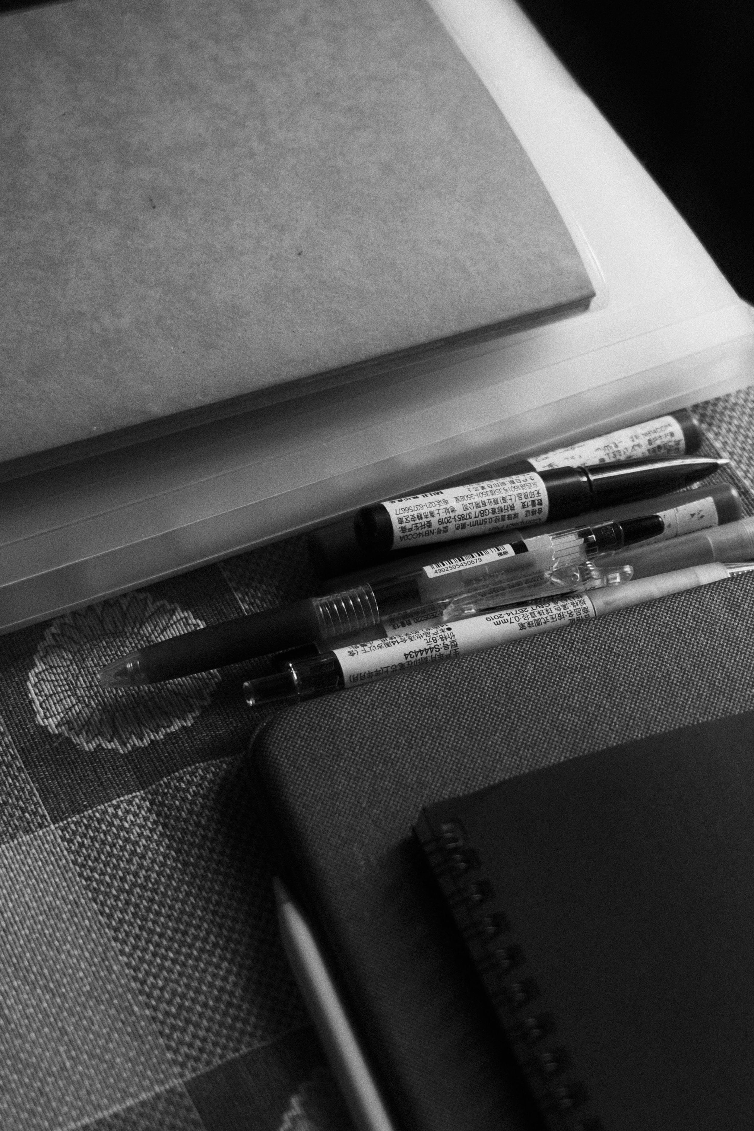 Black and white photo of pens, notebooks, and a folder on a patterned surface.