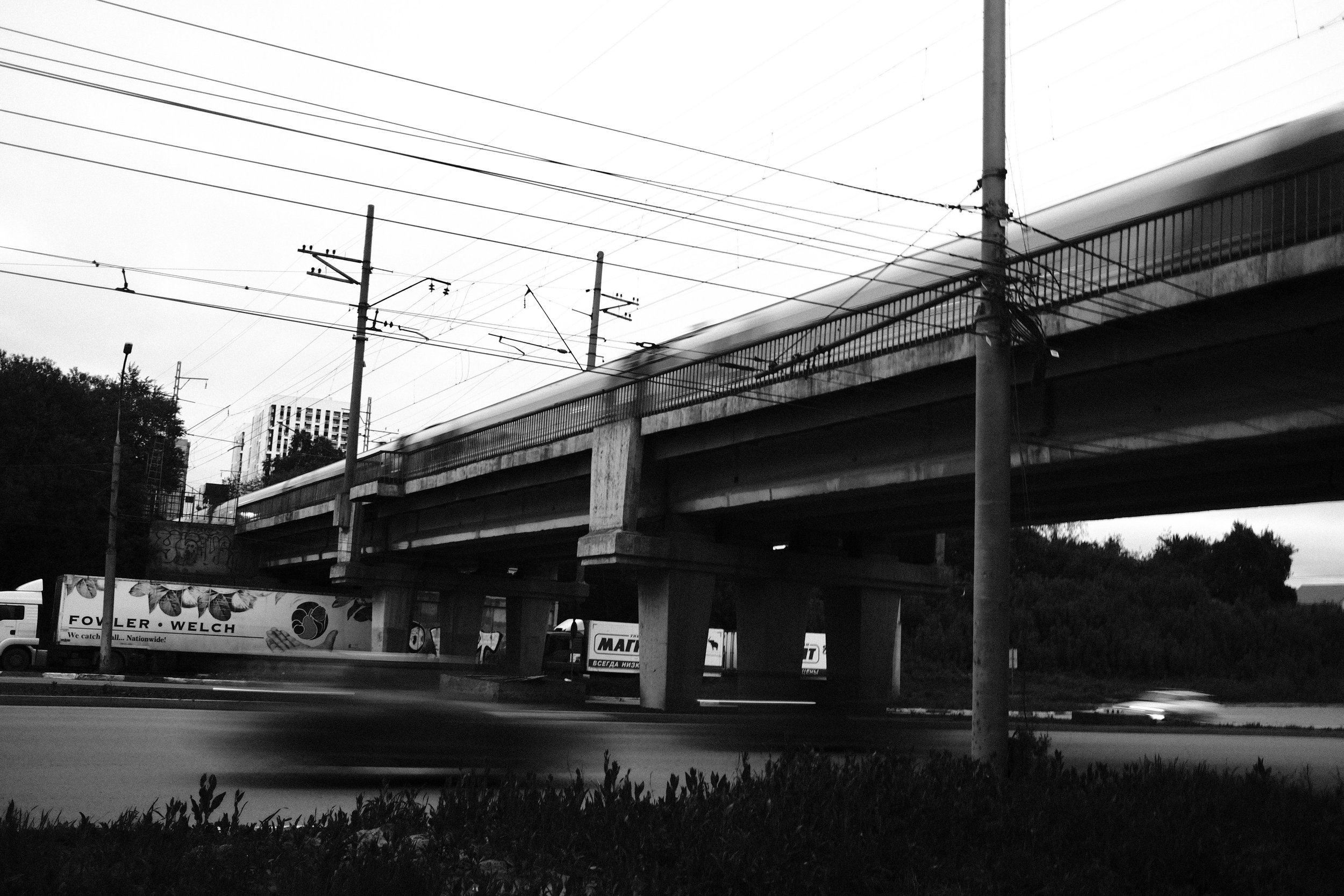 Black and white photo of an overpass with moving vehicles underneath, a truck on the left with advertising, and a blurred car in motion on the road.