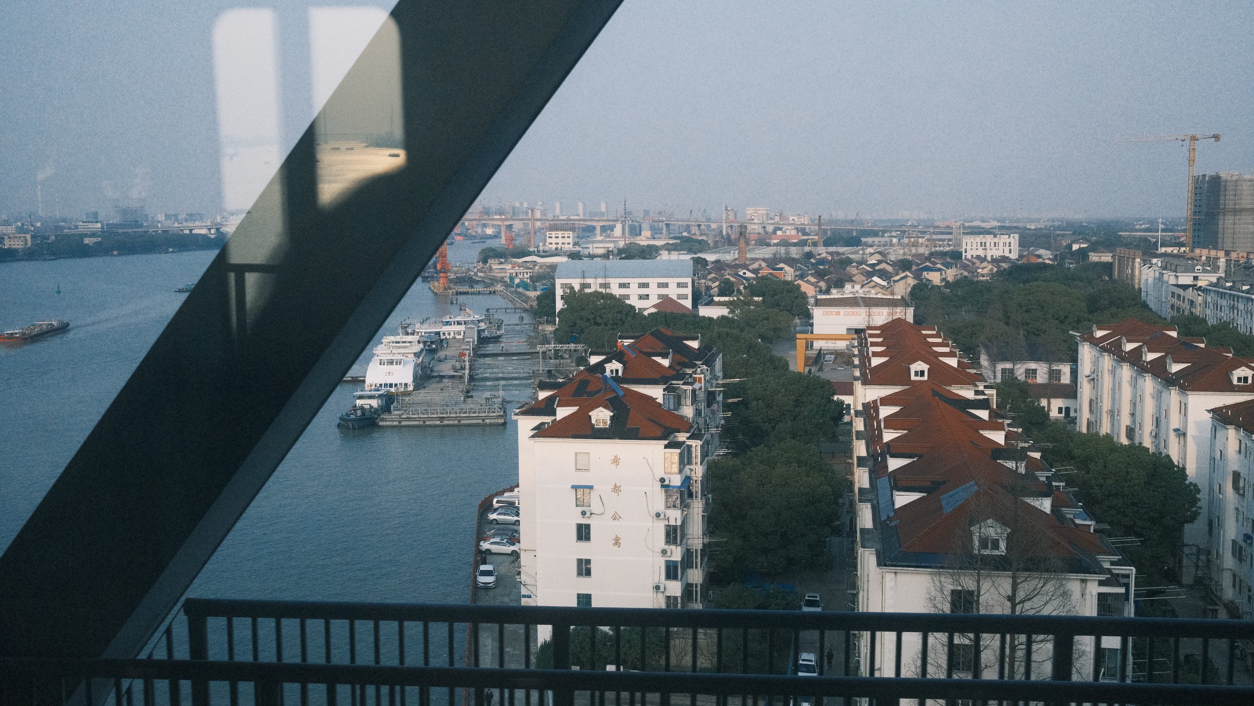 Cityscape view from a high balcony showing waterway, boats, residential buildings with red roofs, and trees, with a distant bridge and construction crane.