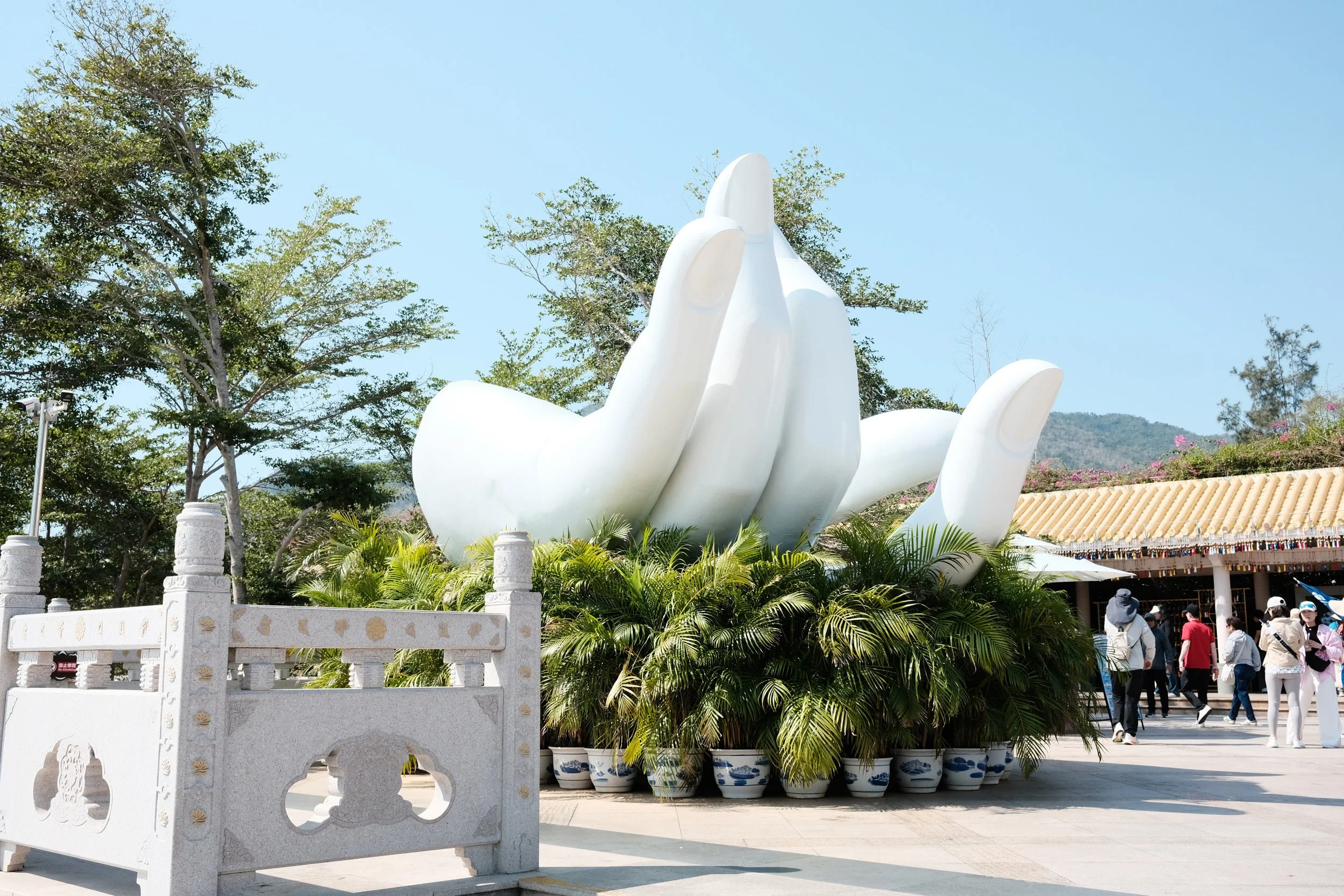 A large white sculpture resembling a hand with fingers together, overlooking a garden of potted green plants, with tall trees and a clear blue sky in the background, and a group of people walking nearby.