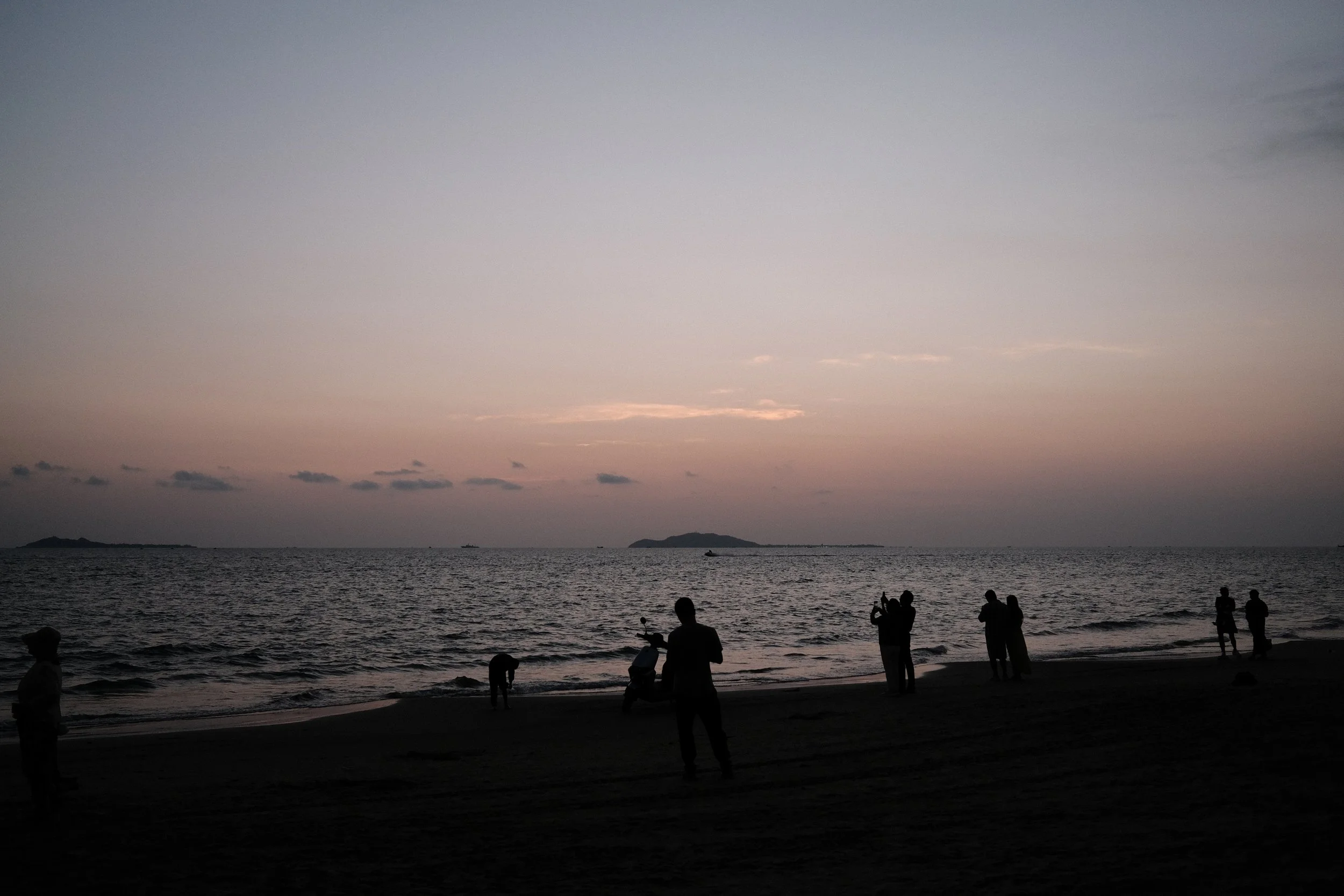 Silhouettes of people on a beach at sunset with the ocean and islands in the background.