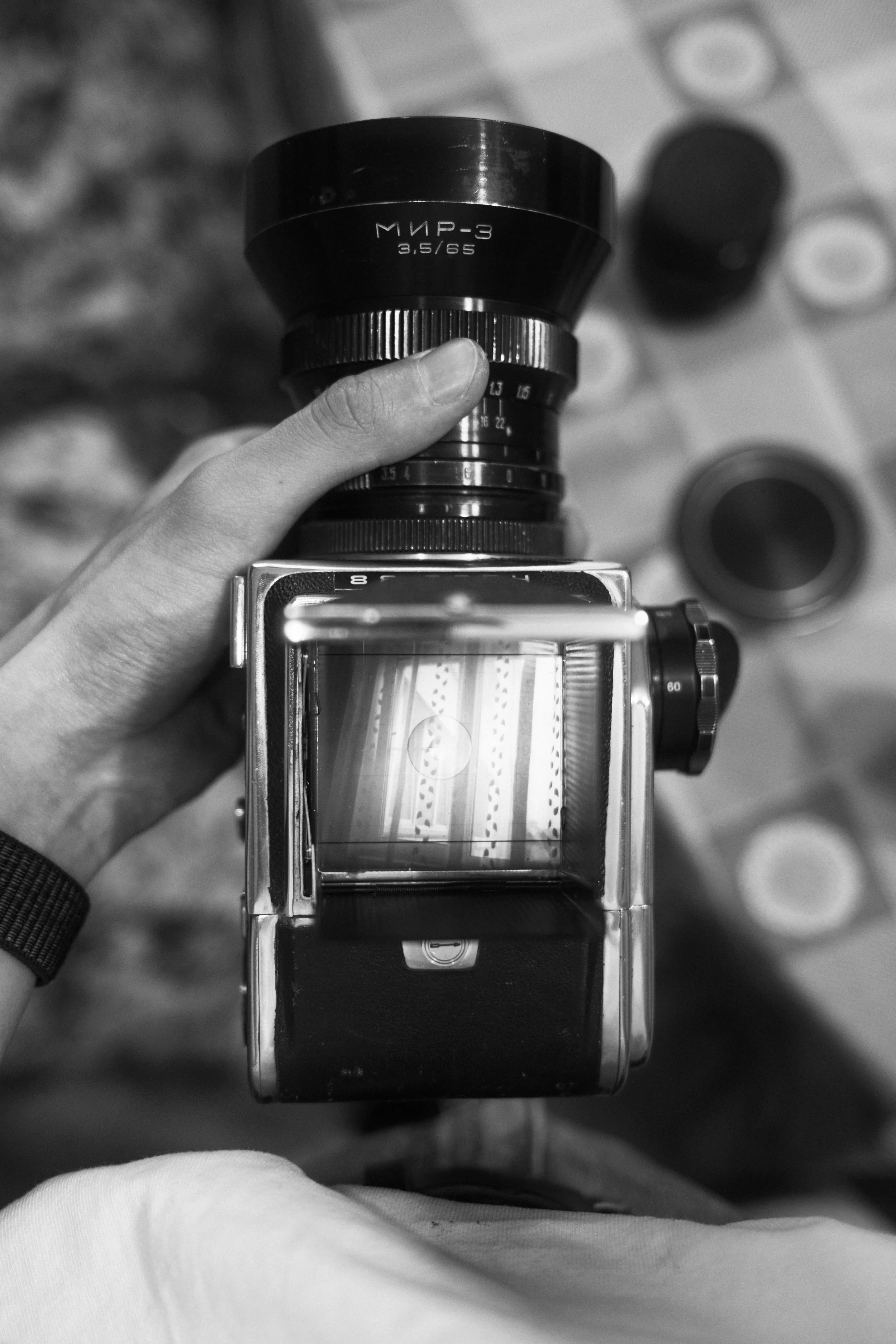Black and white photo of a person holding a vintage film camera with a large lens, viewed from above, with film strips visible through the camera's viewfinder.
