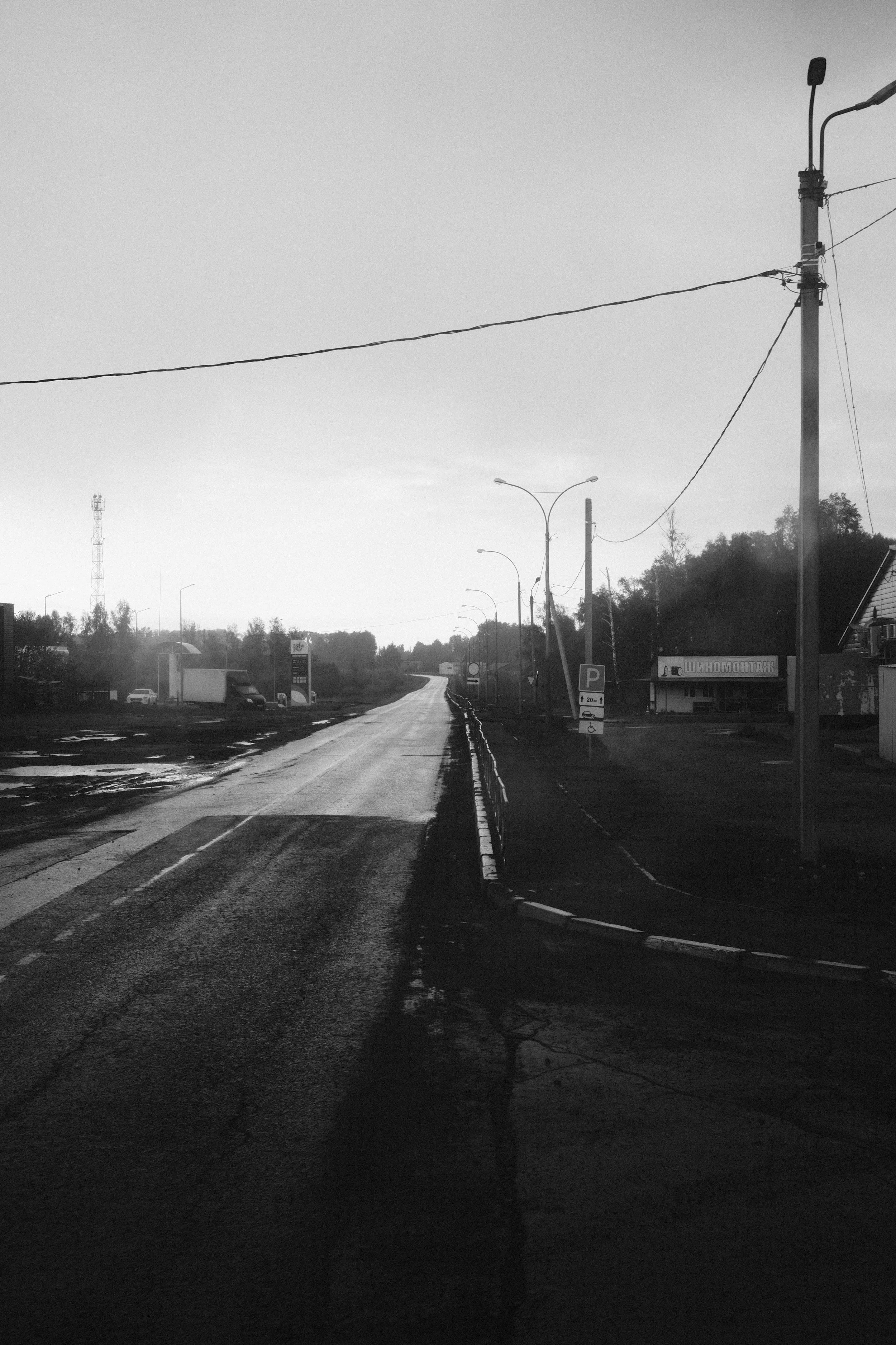 A black and white photo of a rural road extending into the distance with streetlights on its sides, utility poles, and a few small buildings and cars along the roadside.
