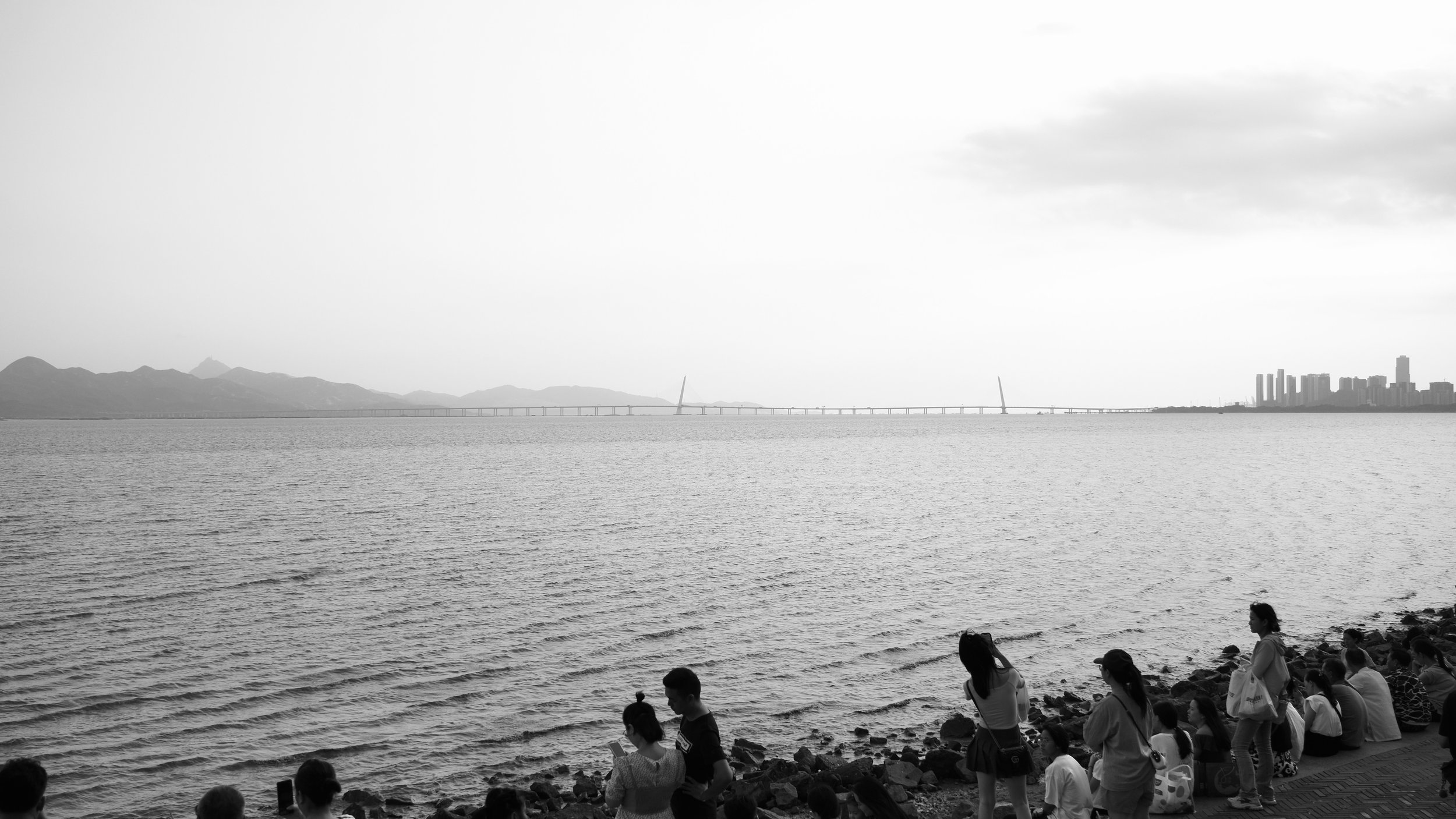 People sitting and standing along a rocky waterfront, overlooking a large body of water with a bridge and distant city skyline in the background.