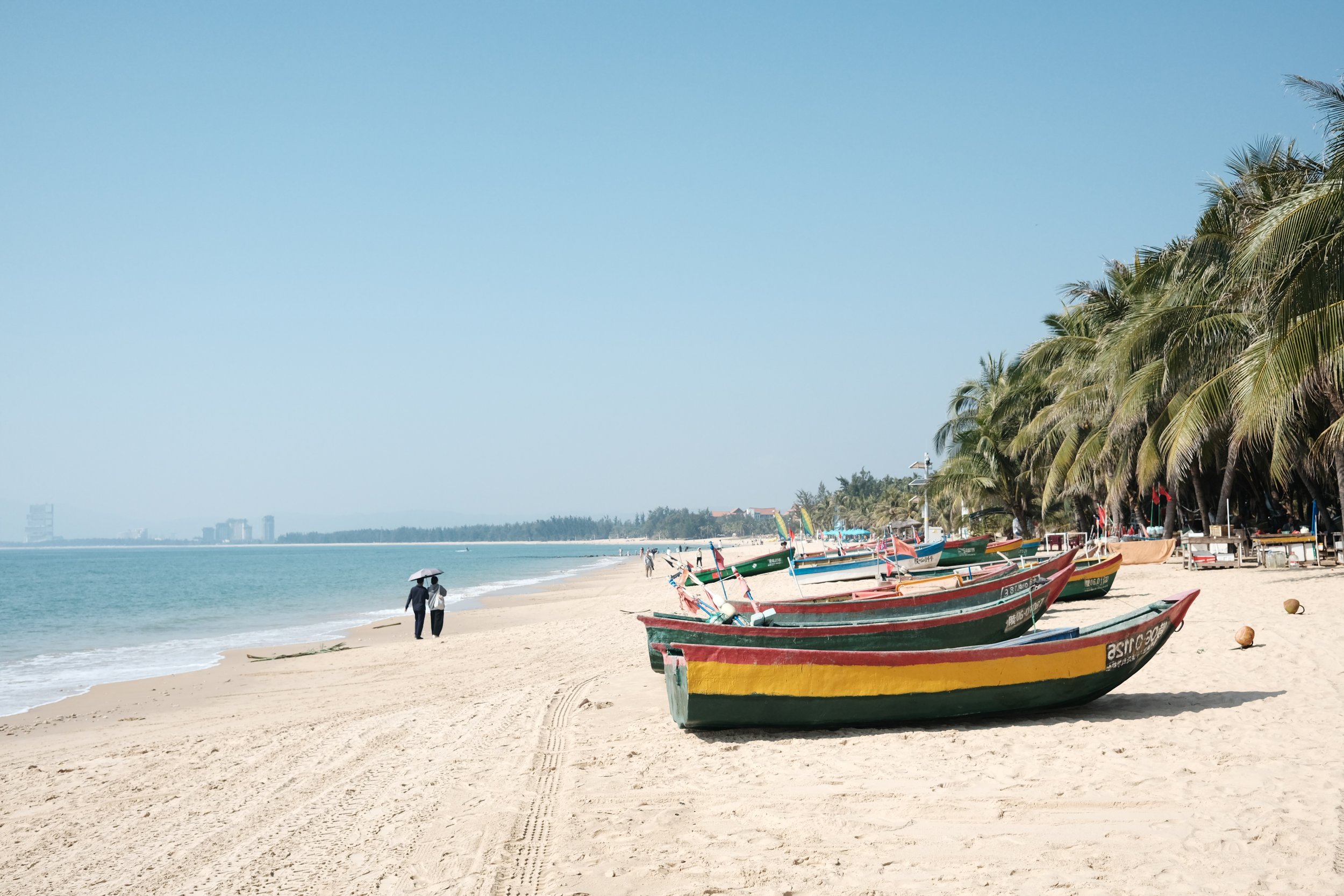 Empty sandy beach with colorful boats and palm trees, two people walking with an umbrella, city skyline in the distance under a clear blue sky.