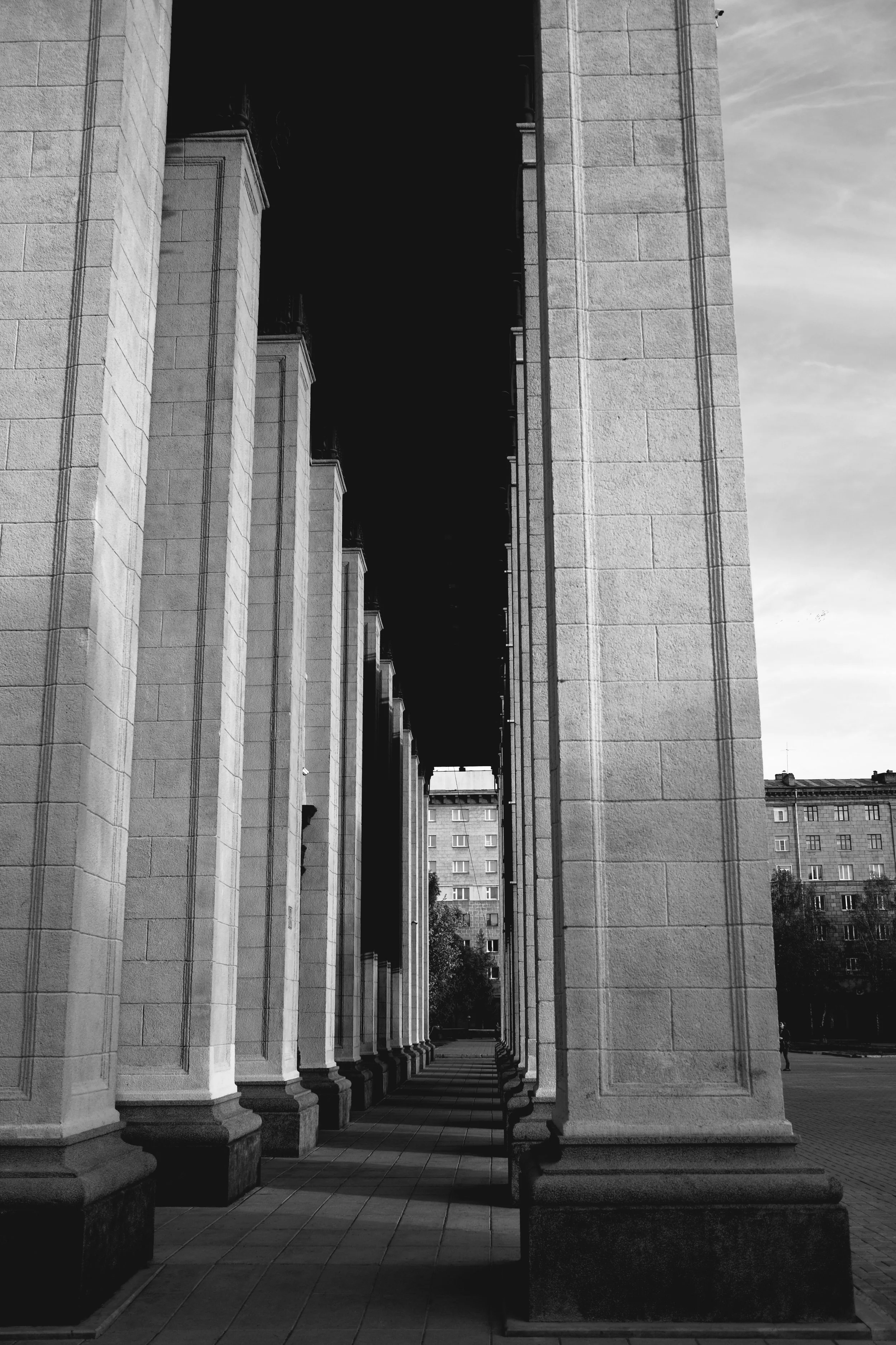Black and white photo of large stone columns in a row, forming a corridor, with a building visible in the distance.