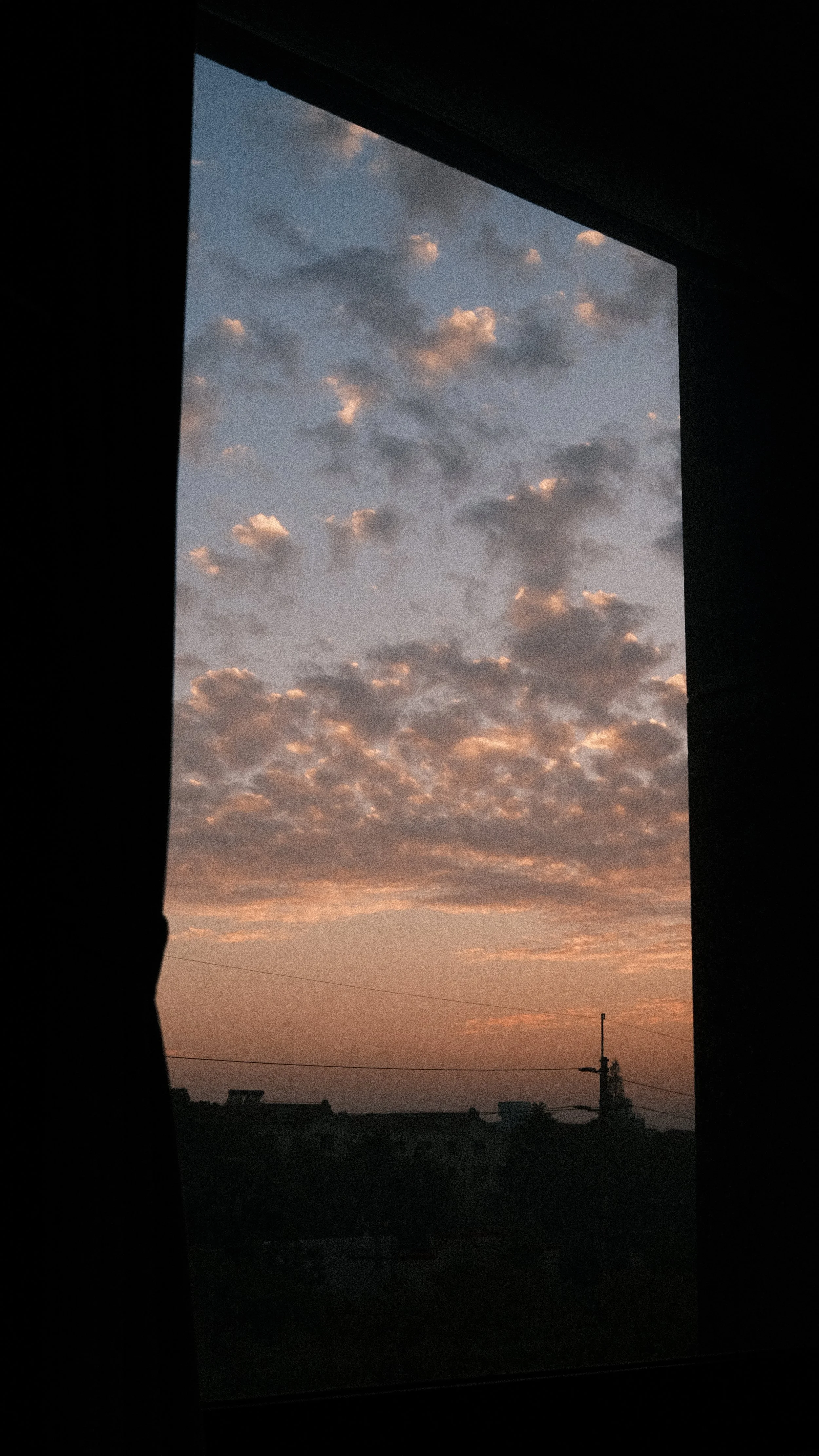 View of a cloudy sunset sky through a window, with dark silhouettes of buildings and power lines in the foreground.