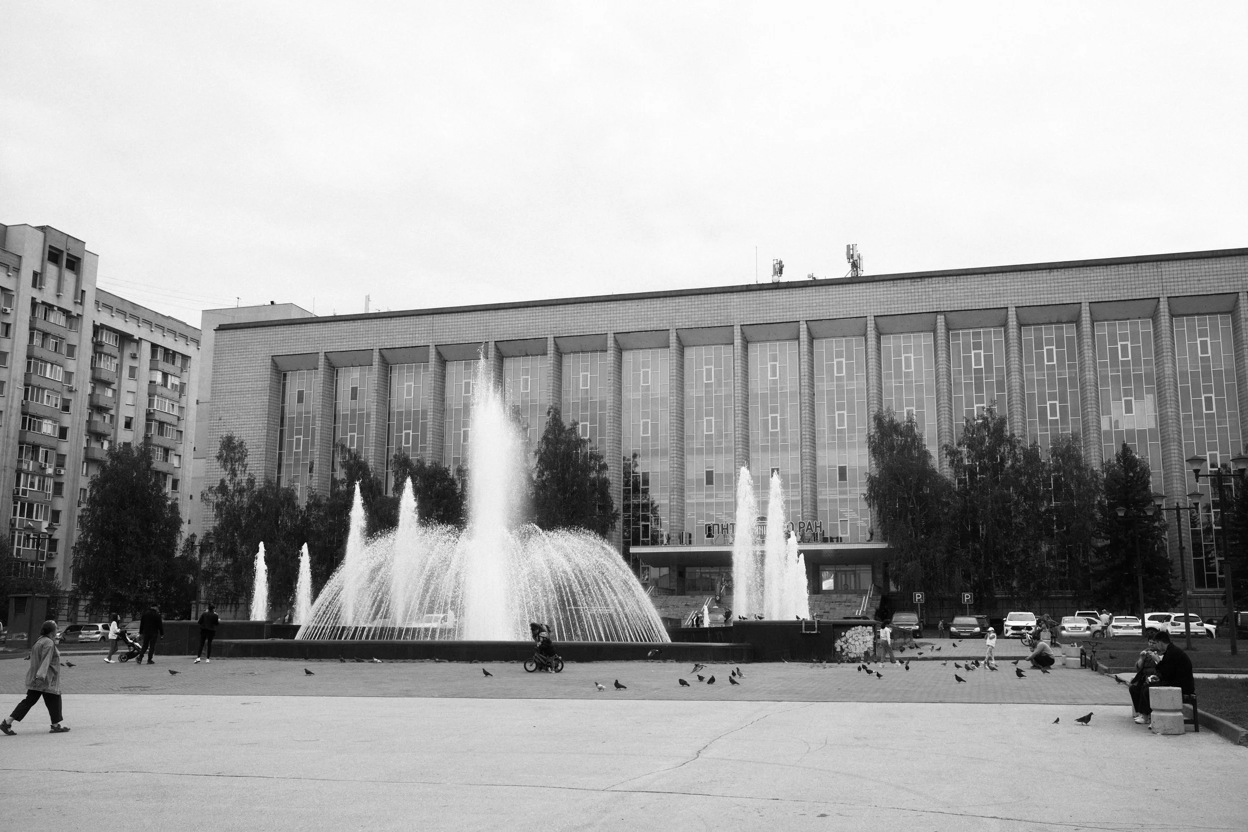 A city square with a large fountain featuring multiple water jets in front of a large modern building with tall windows. Several people are walking, sitting, or riding bicycles around the fountain, and pigeons are scattered on the ground.