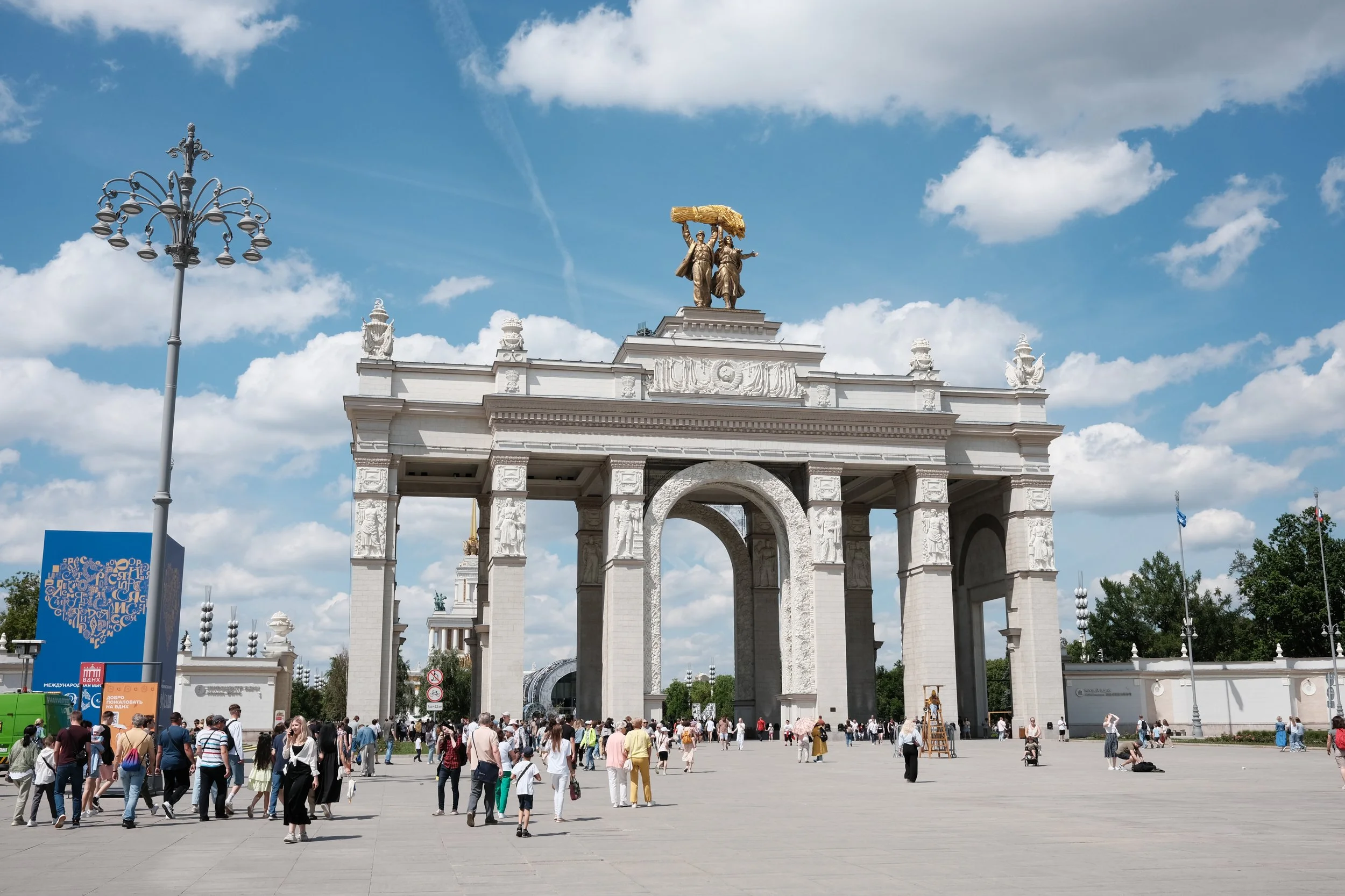 People walking and gathering in front of a large white archway monument with a golden statue of two figures on top, under a partly cloudy sky.