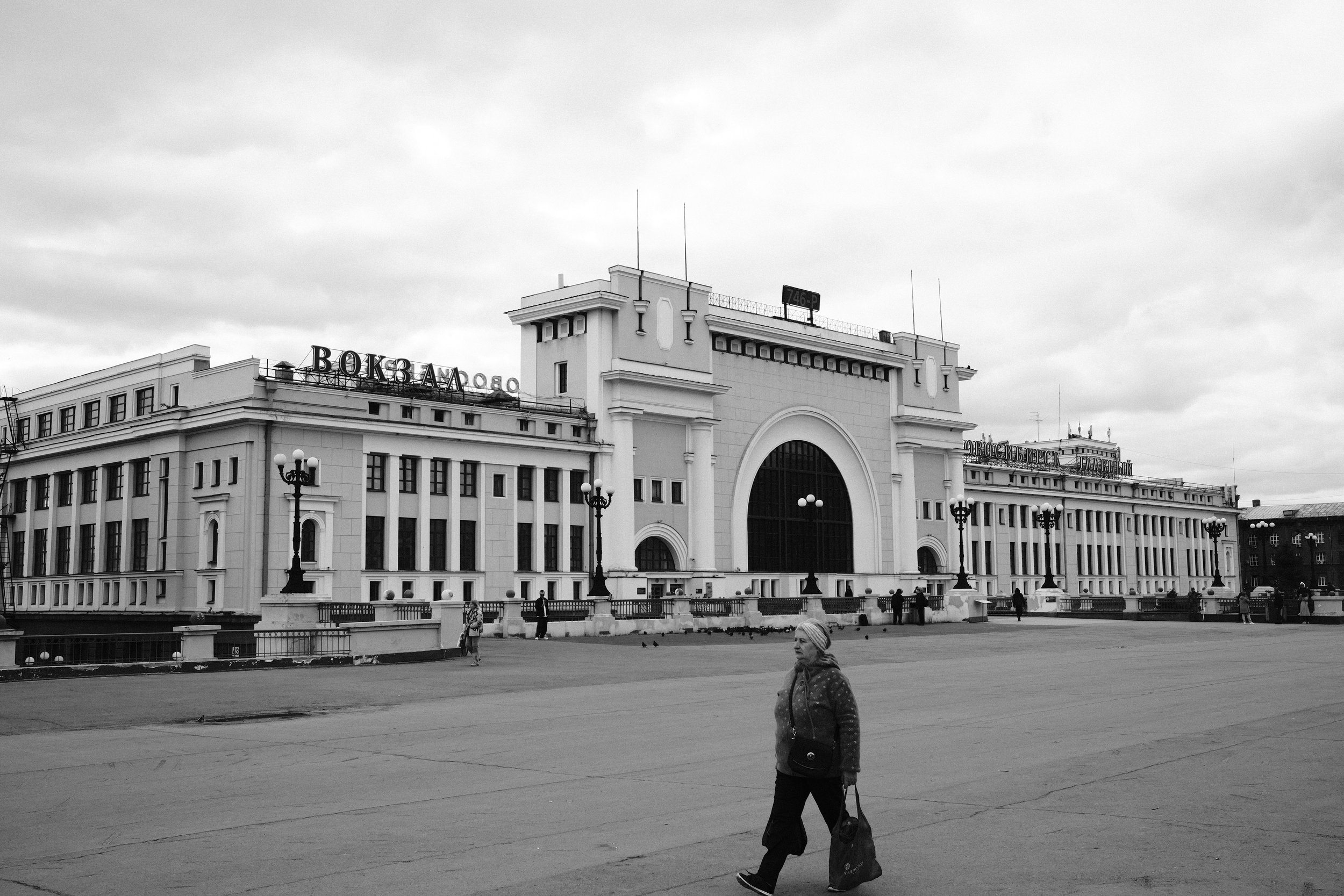 A large historic building with a prominent central arch and multiple windows, located in an open square. There are street lamps, pigeons, and a woman walking with a bag in the foreground. The building has Russian signs on the roof.