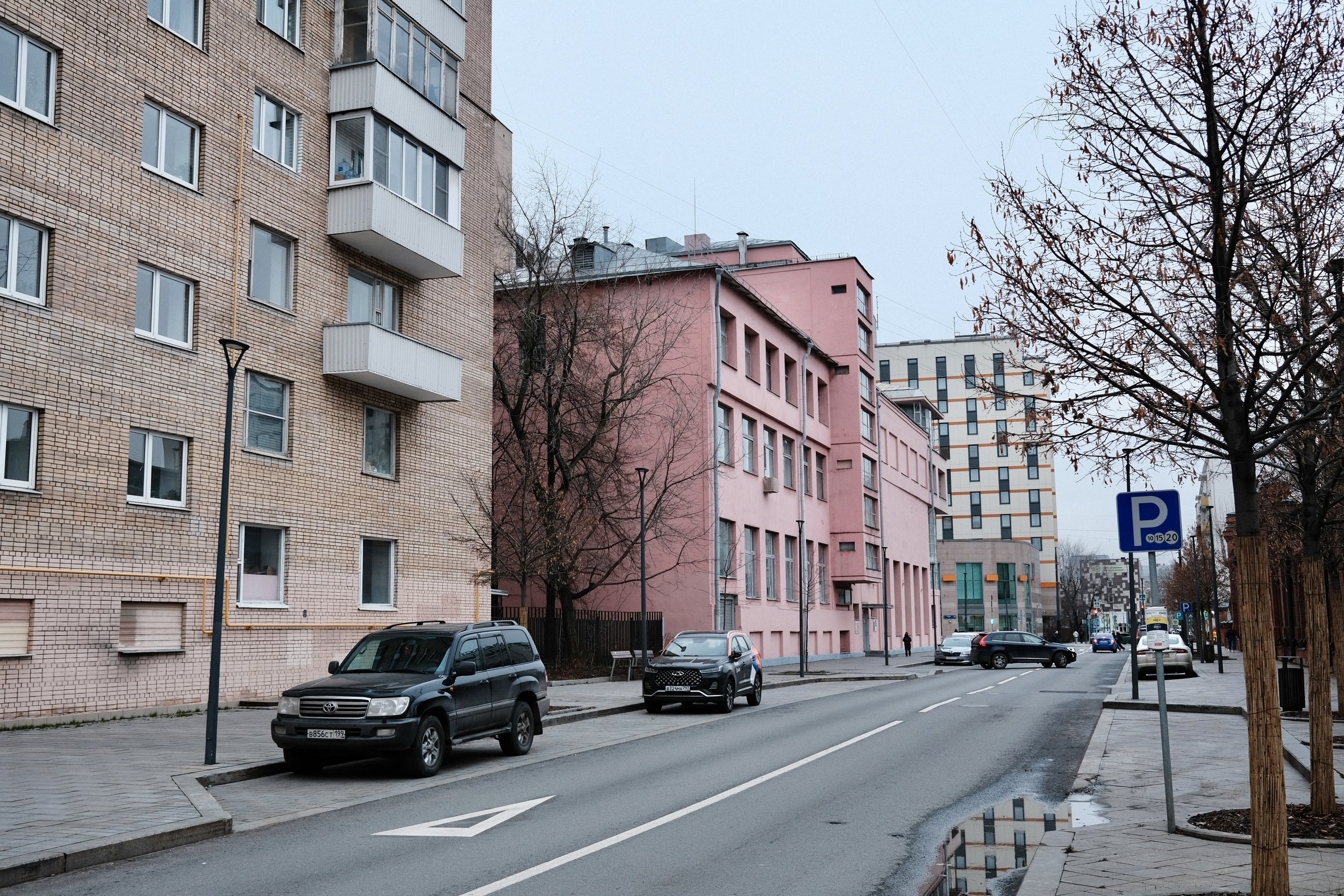 City street scene with parked cars, leafless trees, and multi-story apartment buildings, including a pink building in the center, on an overcast day.