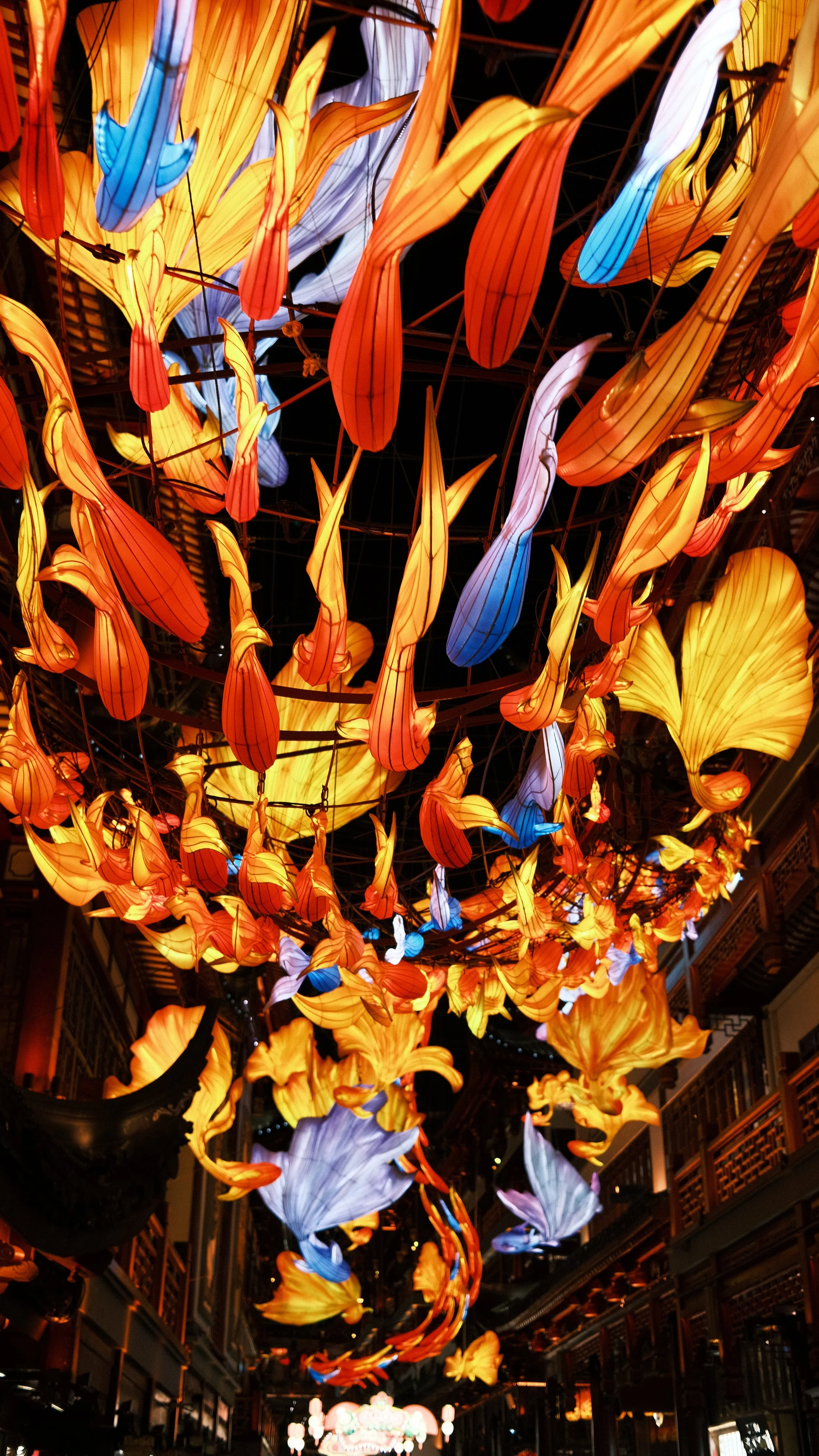 Colorful hanging lanterns resembling flowers and leaves in warm tones of orange, yellow, and some blue and purple shades, illuminating the ceiling of an indoor space.