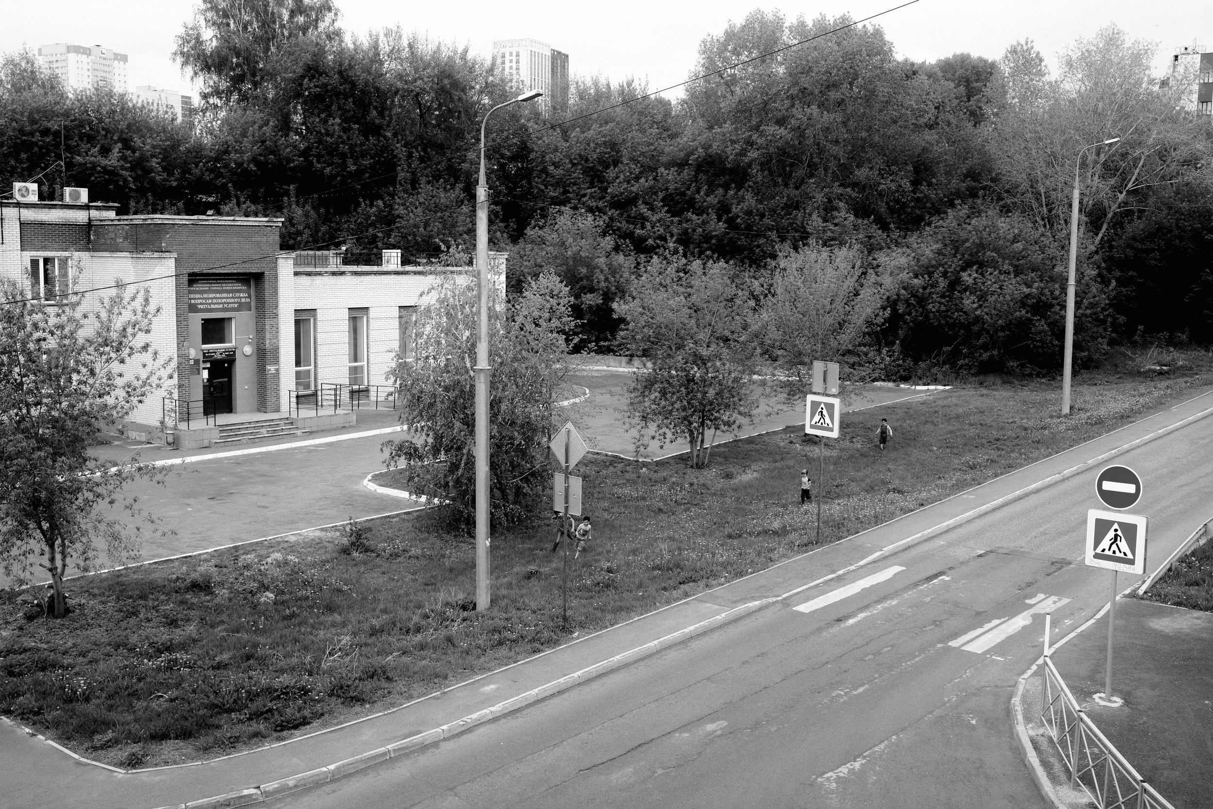 A black and white photograph of a street corner with a small park in the background, featuring trees, a building, street signs including a pedestrian crossing sign and no entry sign, and three children walking in the park.