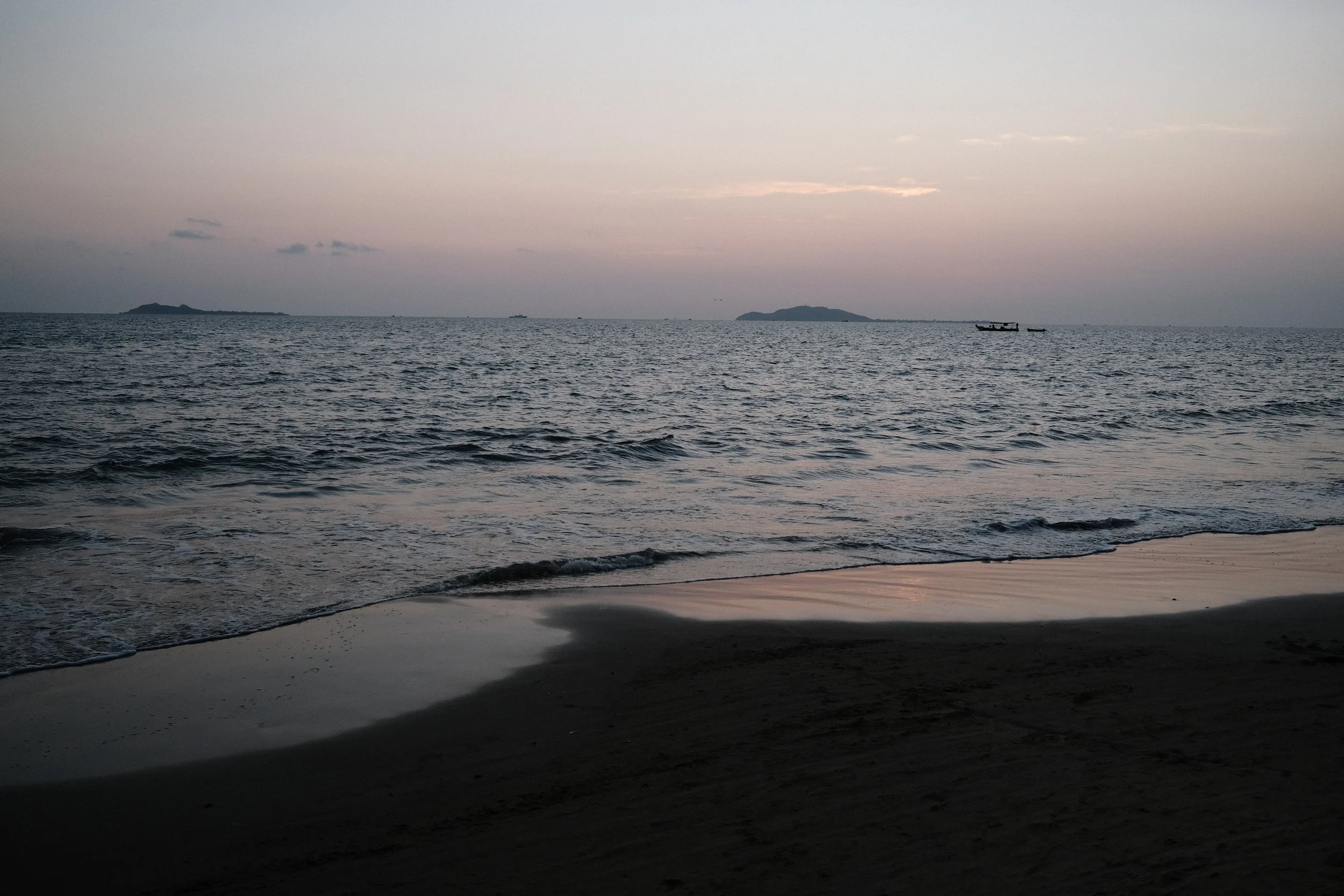 Sunset over the ocean with calm waves, a sandy beach in the foreground, and distant islands and boats on the horizon.