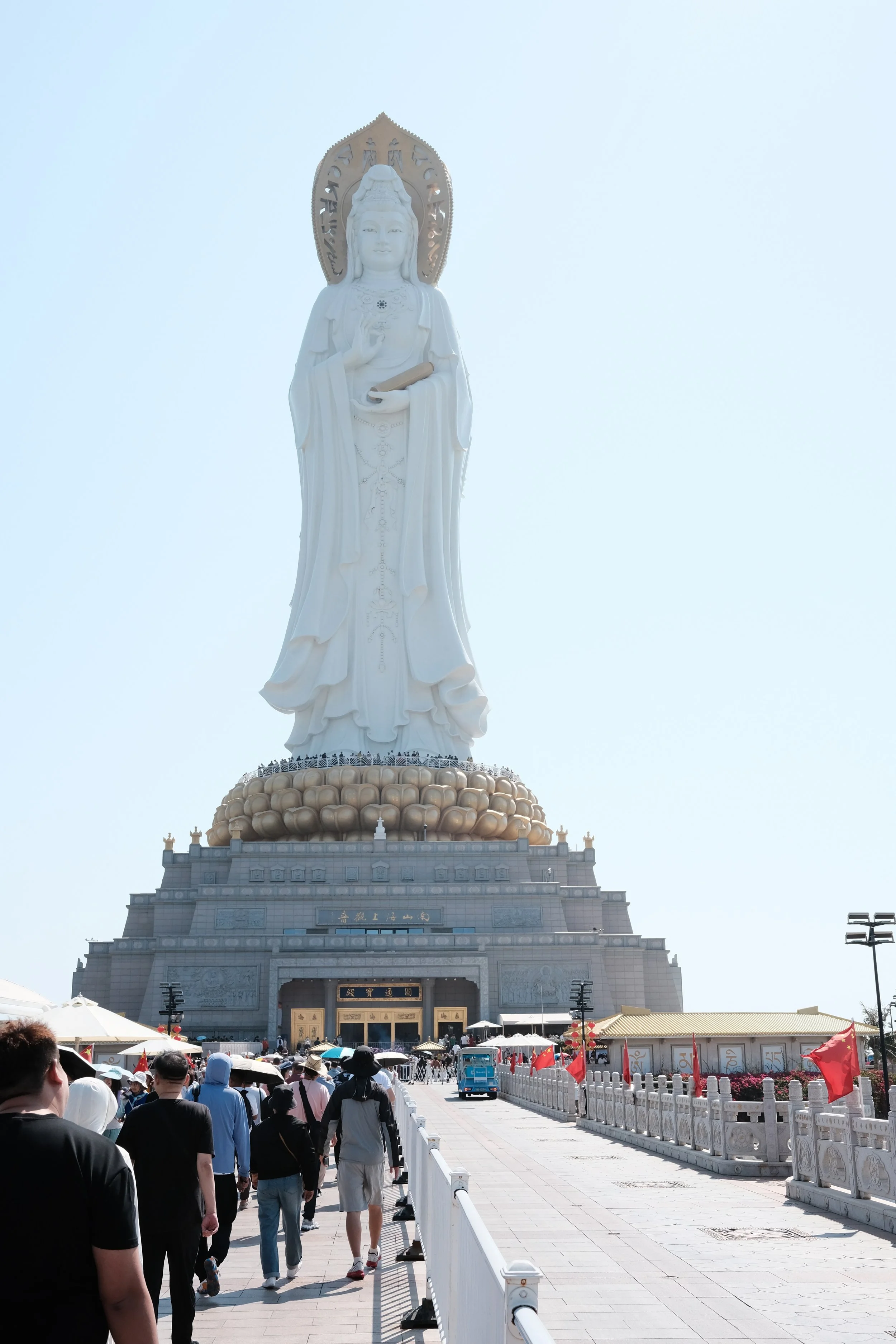 Large white statue of Guanyin Bodhisattva with a gold halo and a scroll, surrounded by a crowd of visitors walking towards a temple entrance on a bright day.