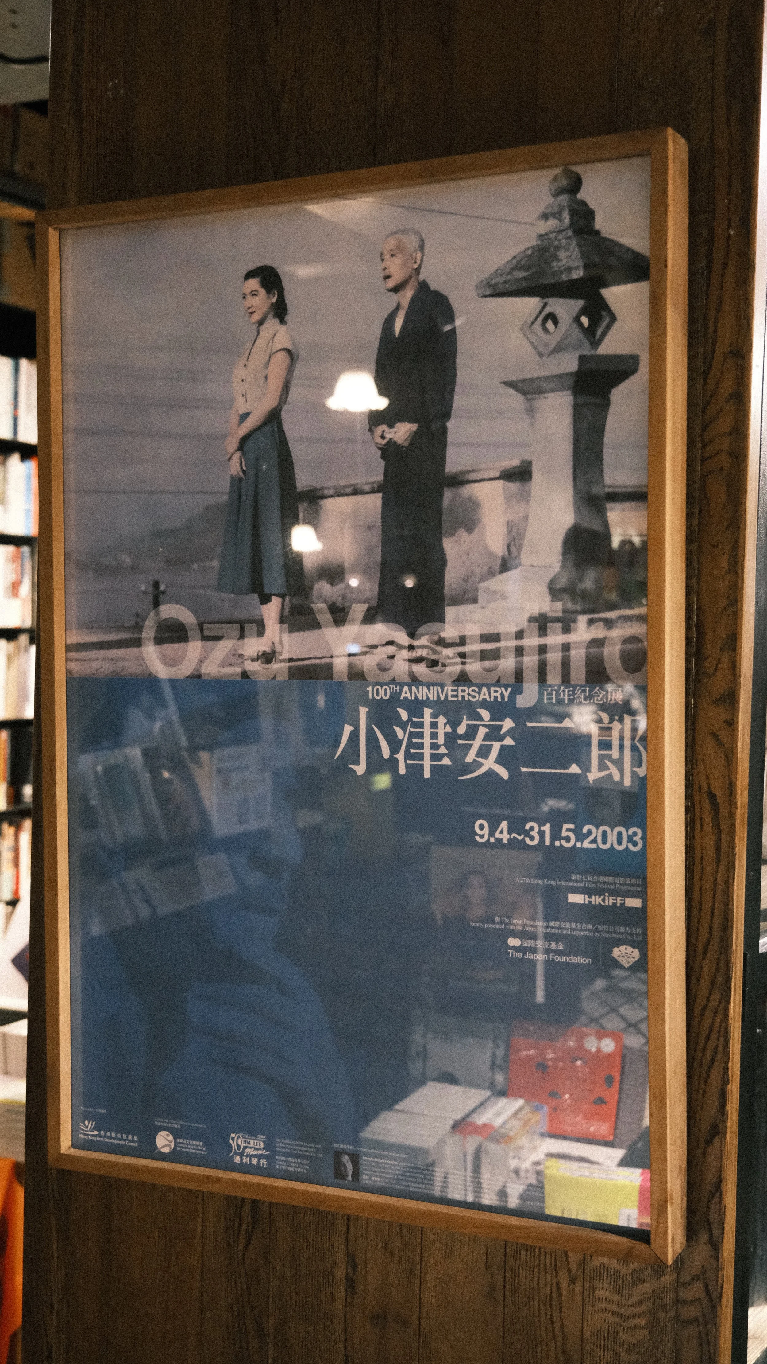A poster commemorating the 100th anniversary of Ozamu Tezuka featuring a black-and-white photograph of a man and woman standing outdoors, with Japanese text and event details below.