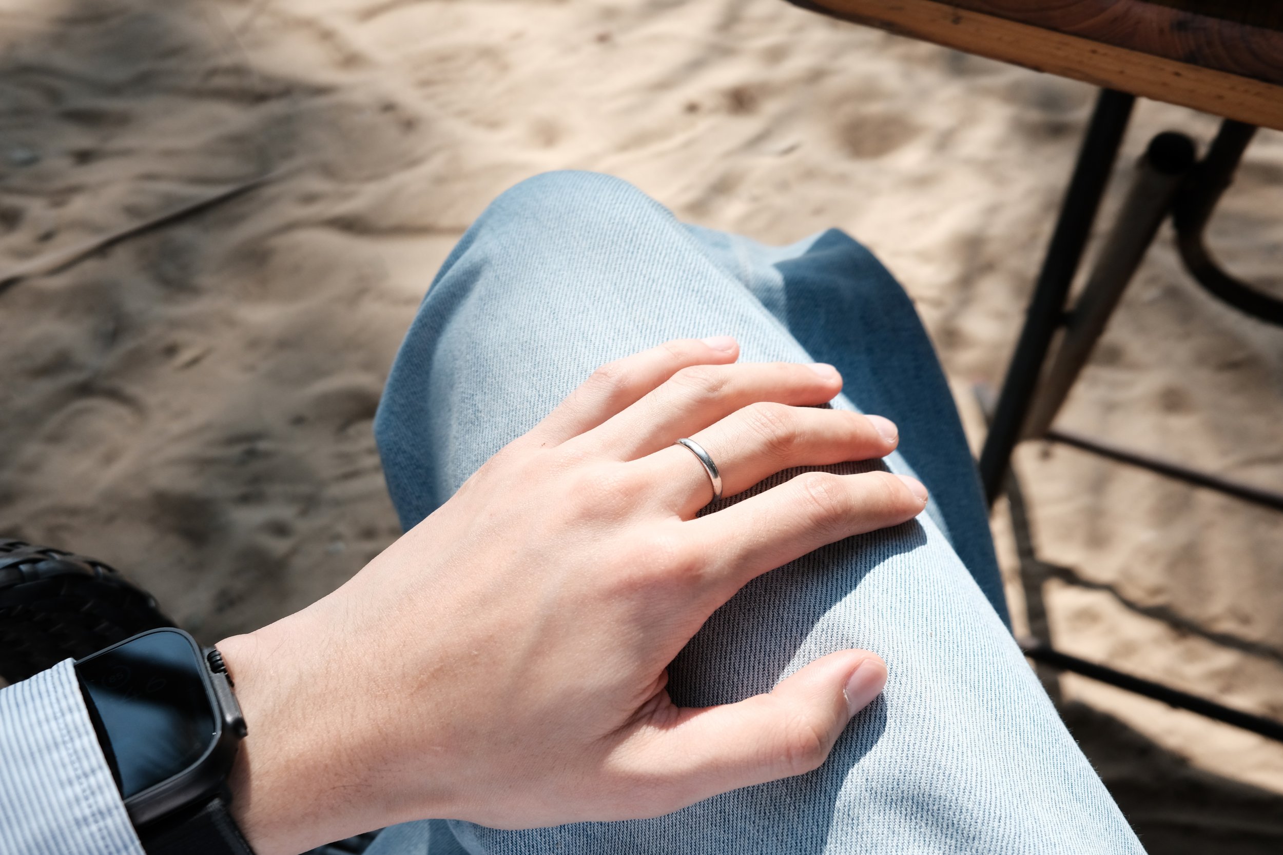 A person's hand with a silver ring resting on their leg, which is covered in light blue denim jeans. The person is sitting outdoors on sandy ground with a wooden surface nearby.