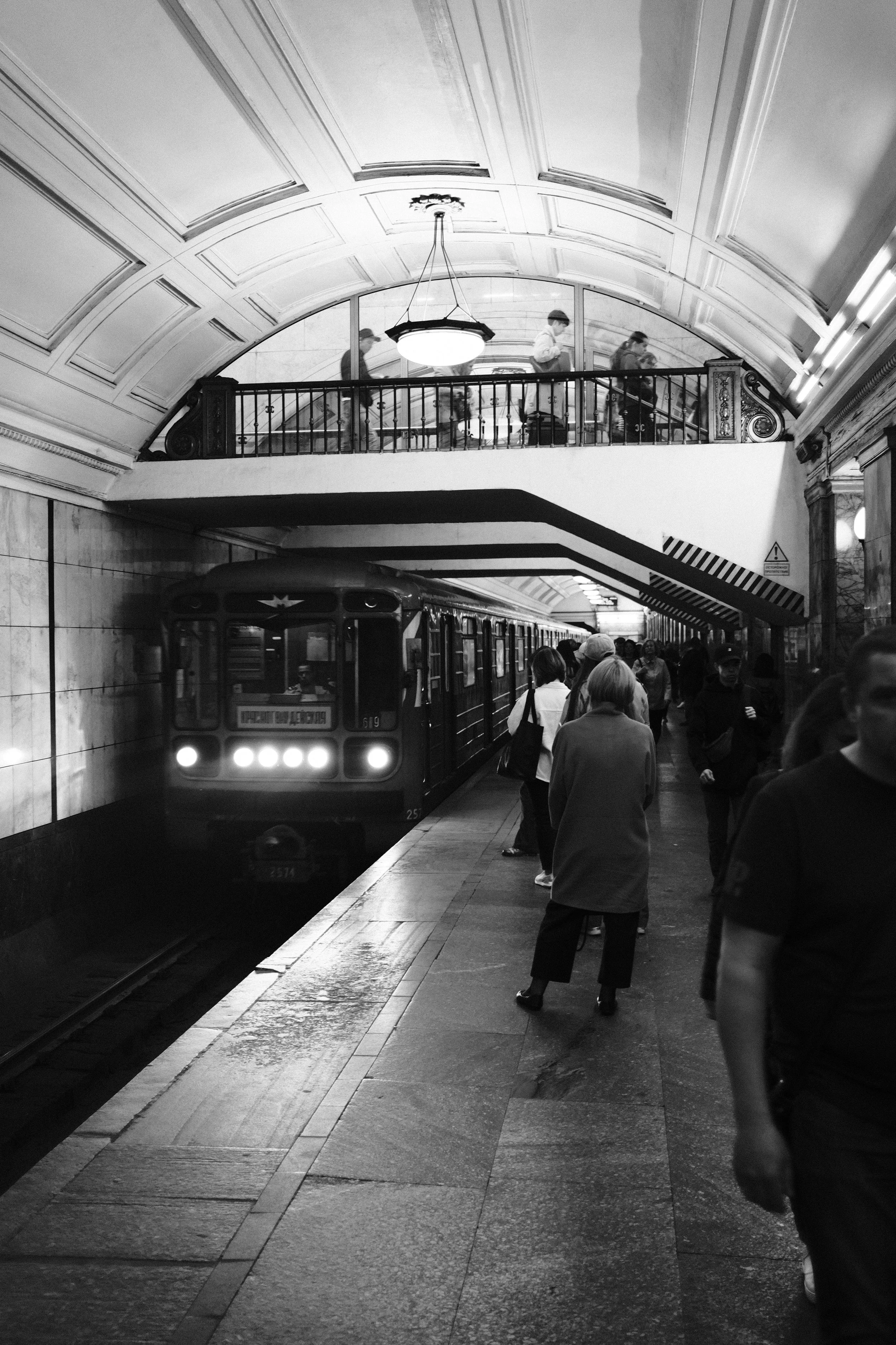 Black and white photo of a subway station with a train arriving and people waiting on the platform. An upper level with few people walking and a large ornate ceiling light.
