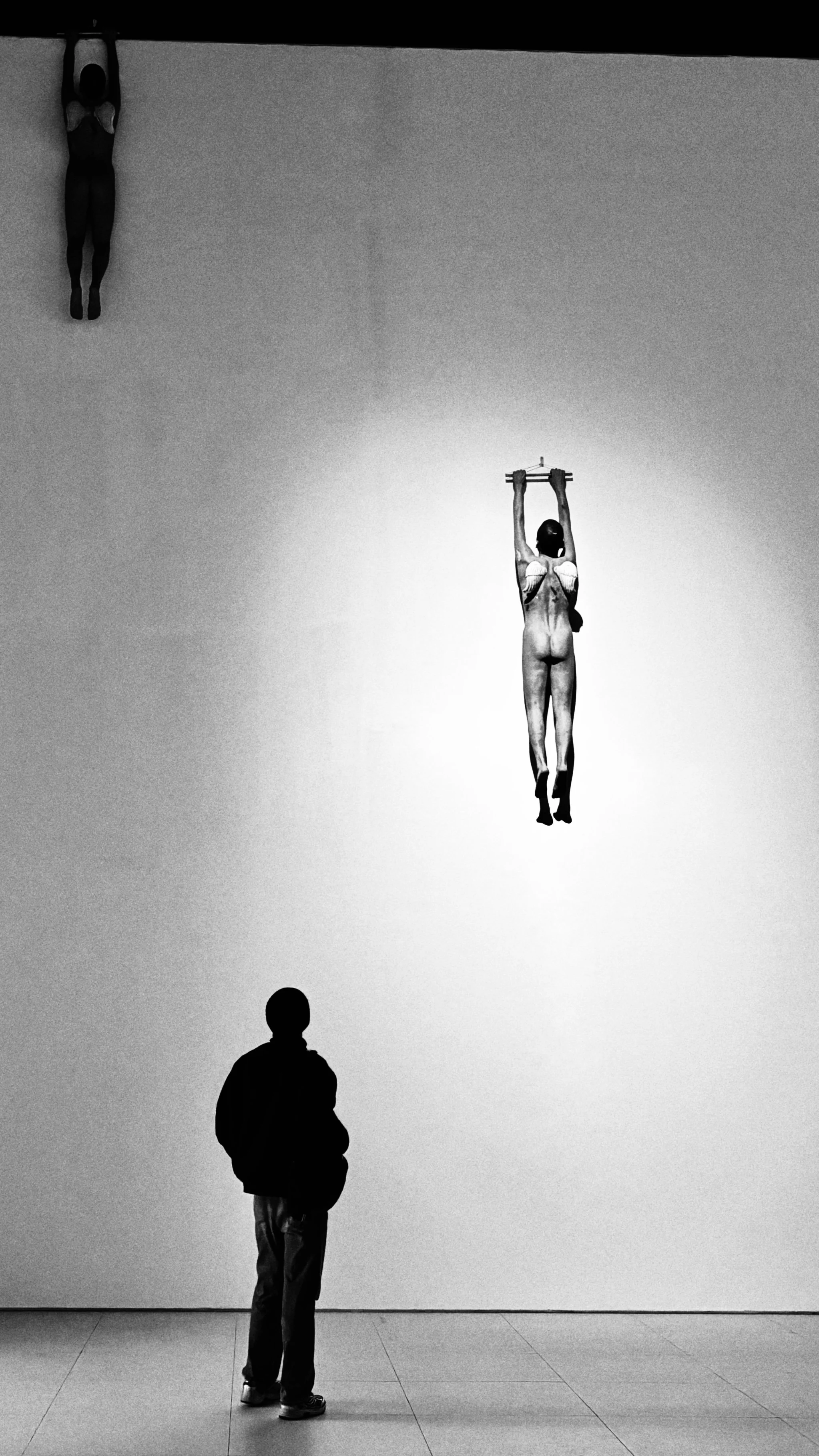 A person stands and observes a black-and-white art installation of three hanging female figures, with one figure in the center holding a bar above her head.