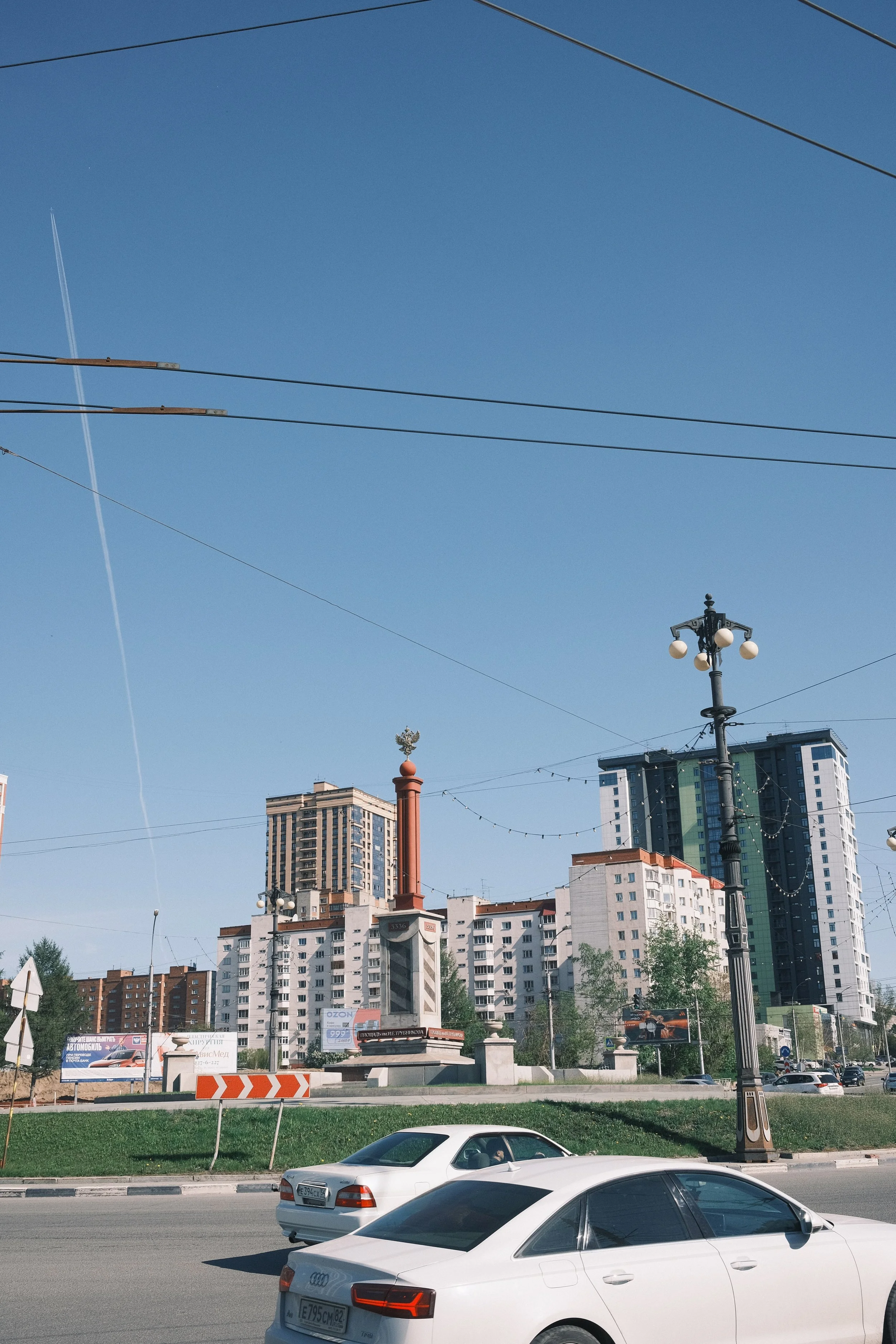 City street with cars, streetlights, and a monument, with high-rise buildings in the background under a clear blue sky.