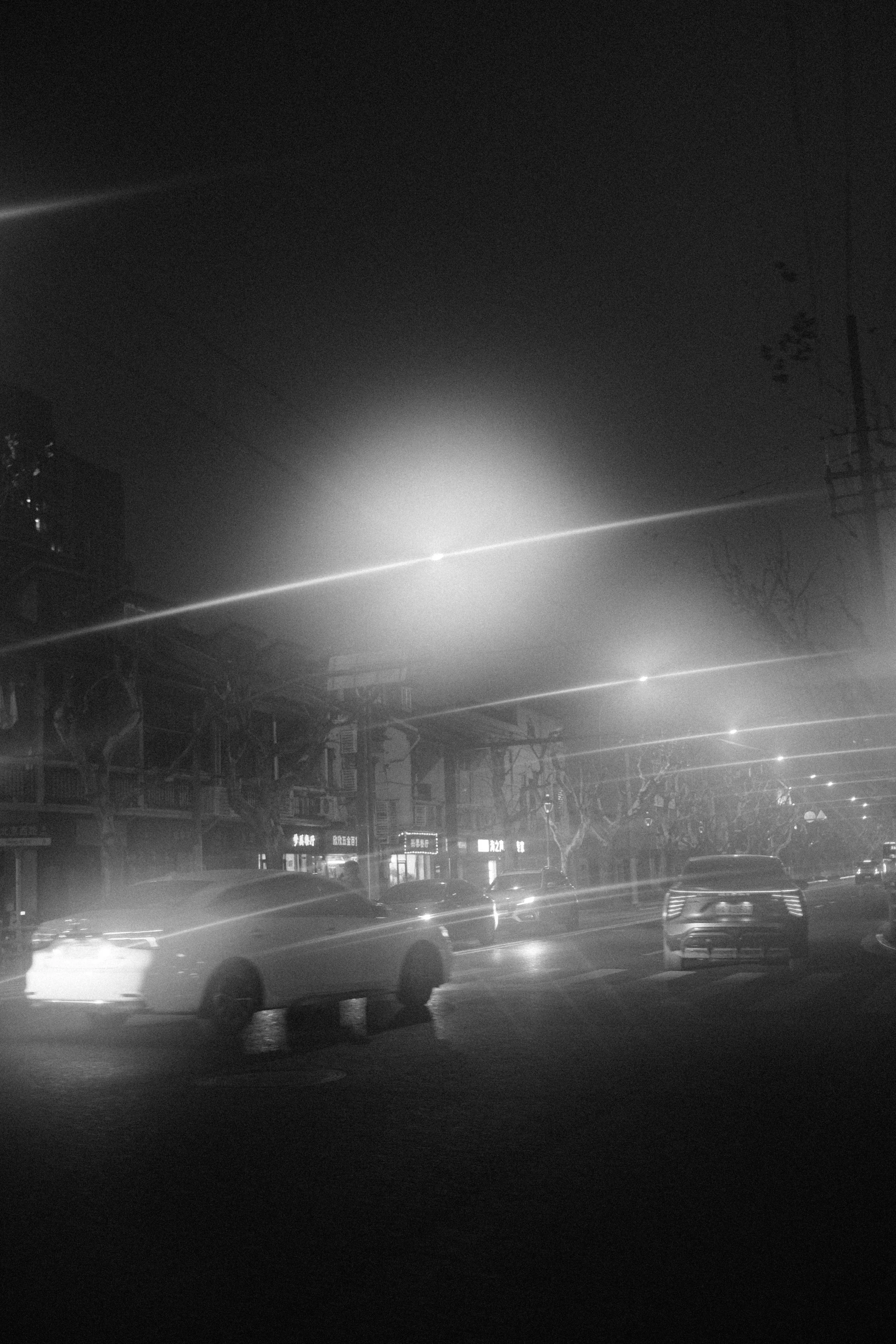 A black and white photo of a nighttime city street with cars parked and on the move, bright streetlights, trees lining the sidewalk, and buildings in the background.