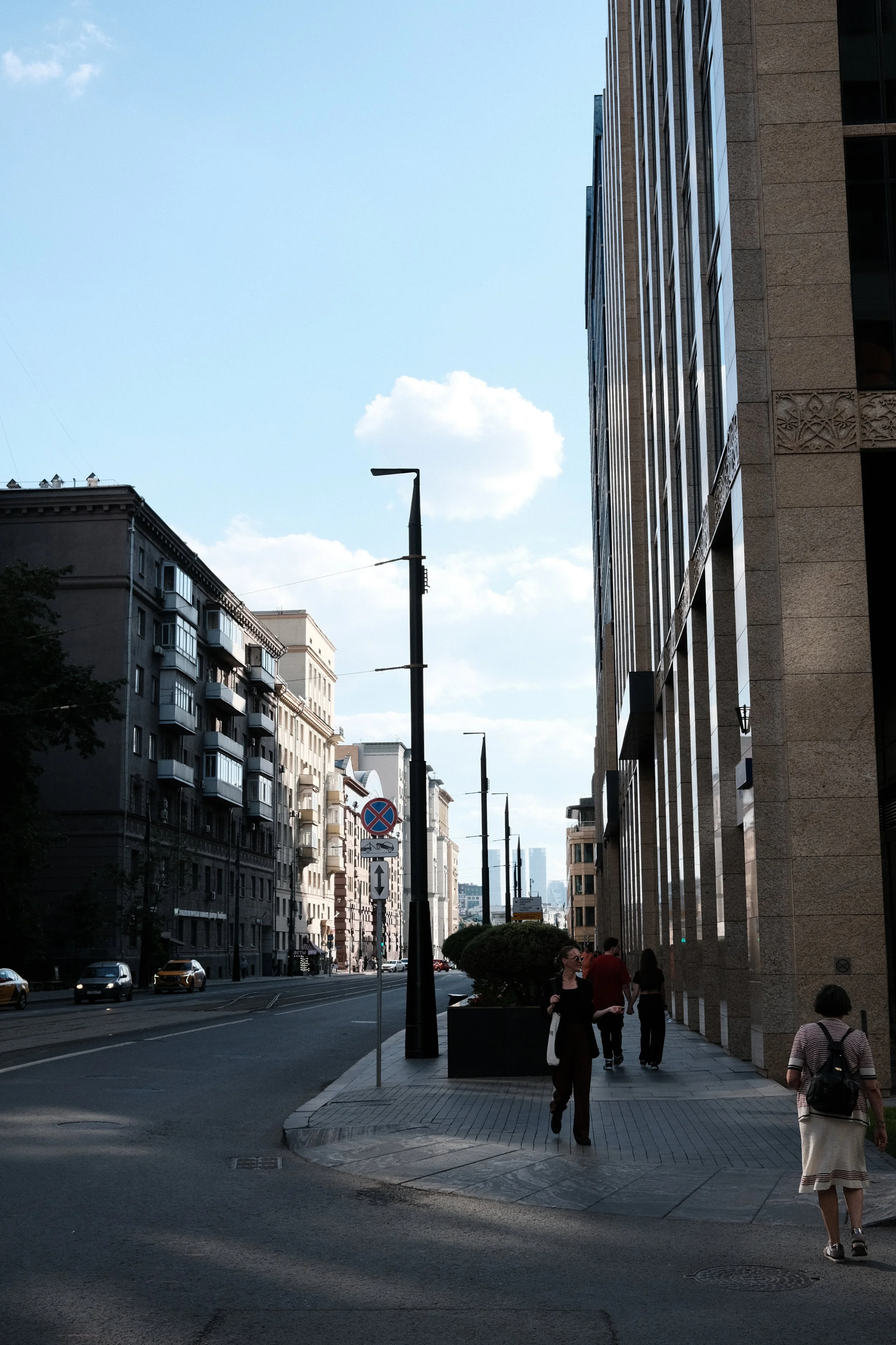 Urban city street scene with tall buildings, sidewalks, street lamps, pedestrians, and cars under a partly cloudy sky.