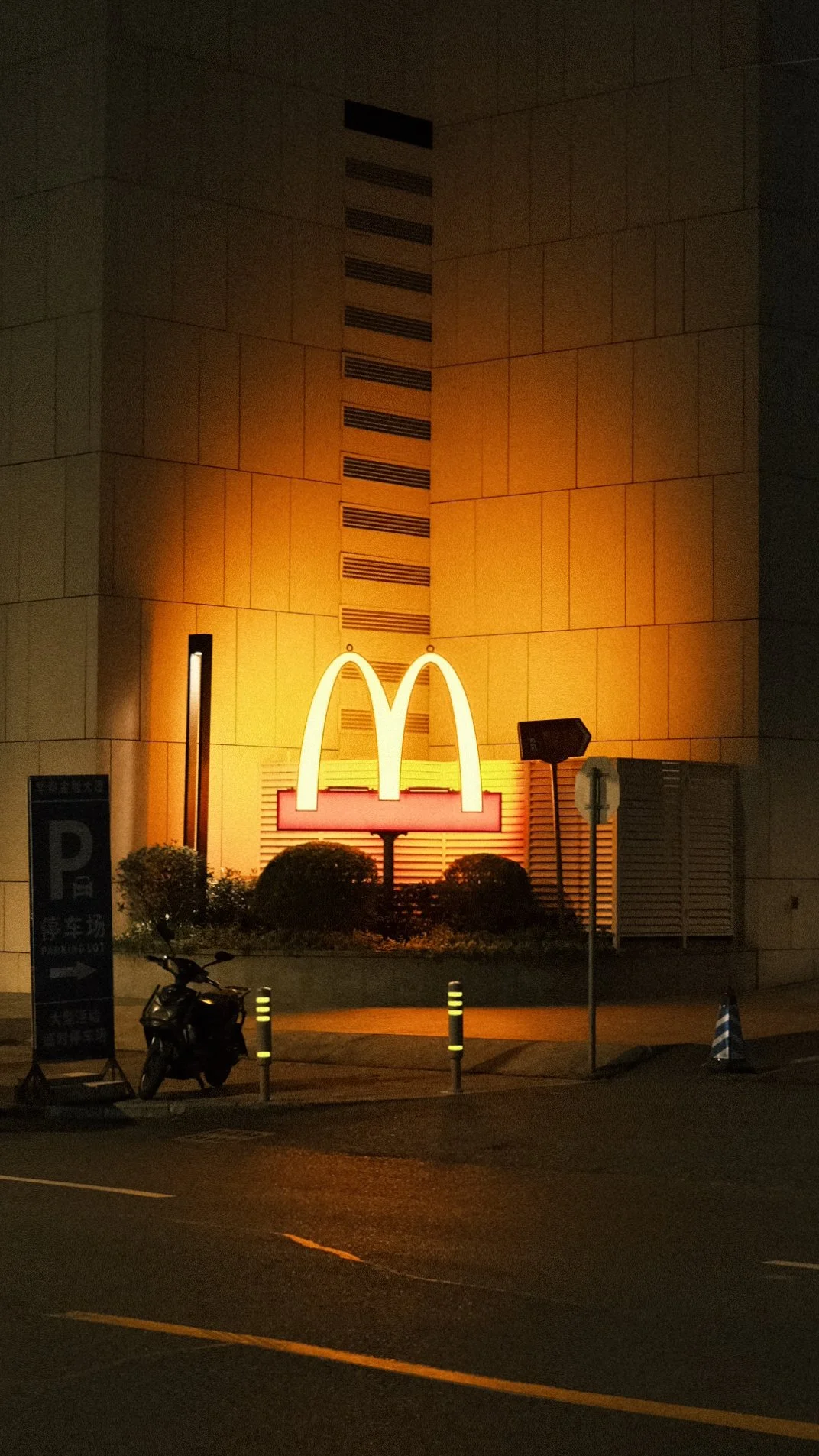 Nighttime photo of a McDonald's sign with the iconic yellow arches on a building with a yellowish-orange exterior. There are parking signs, a scooter, and some small bushes in front.