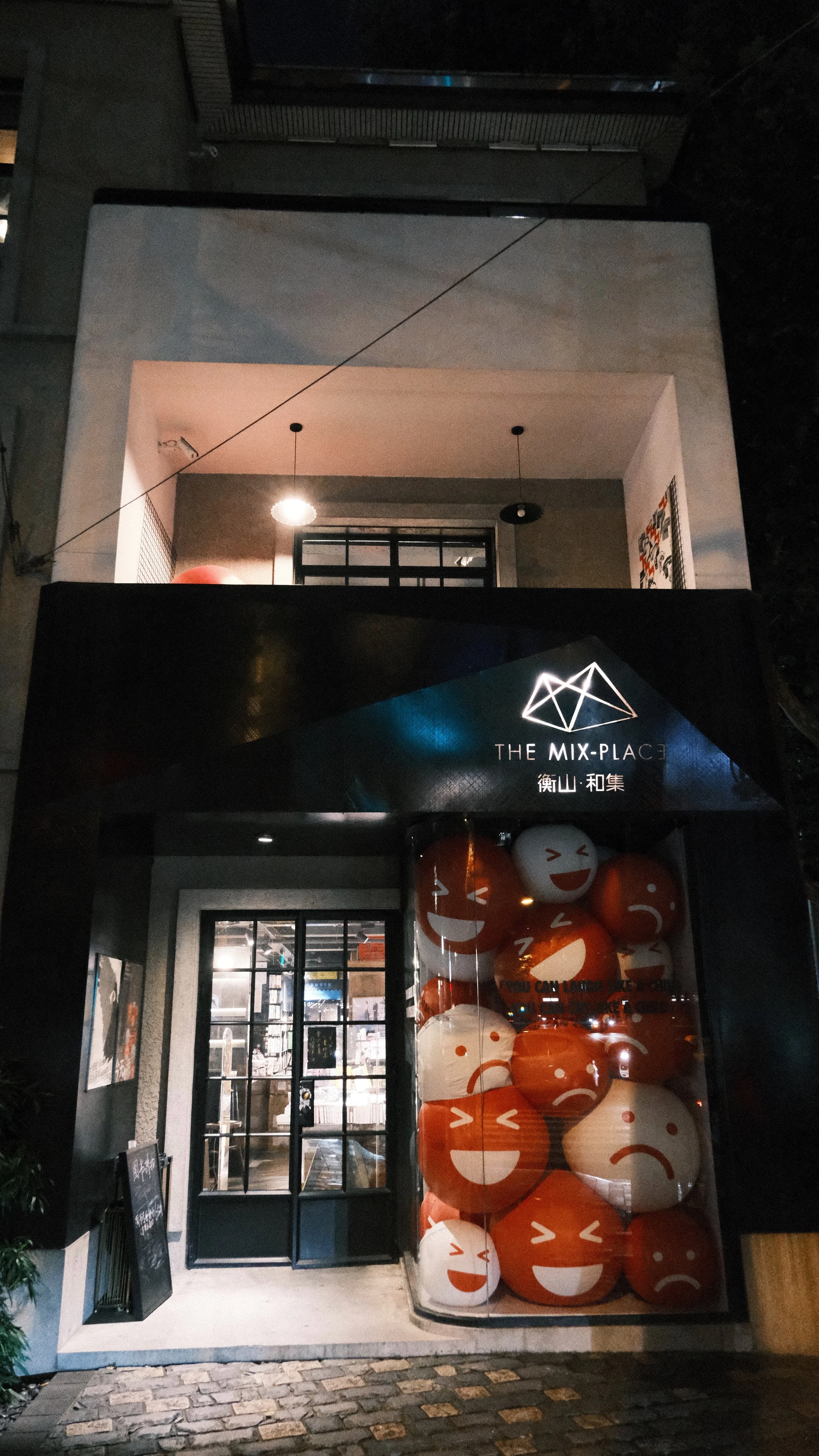 Night view of a modern building with a sign that reads 'THE MIX-PLACE' and display of large orange and white emoji faces with various expressions in a glass case at the entrance.