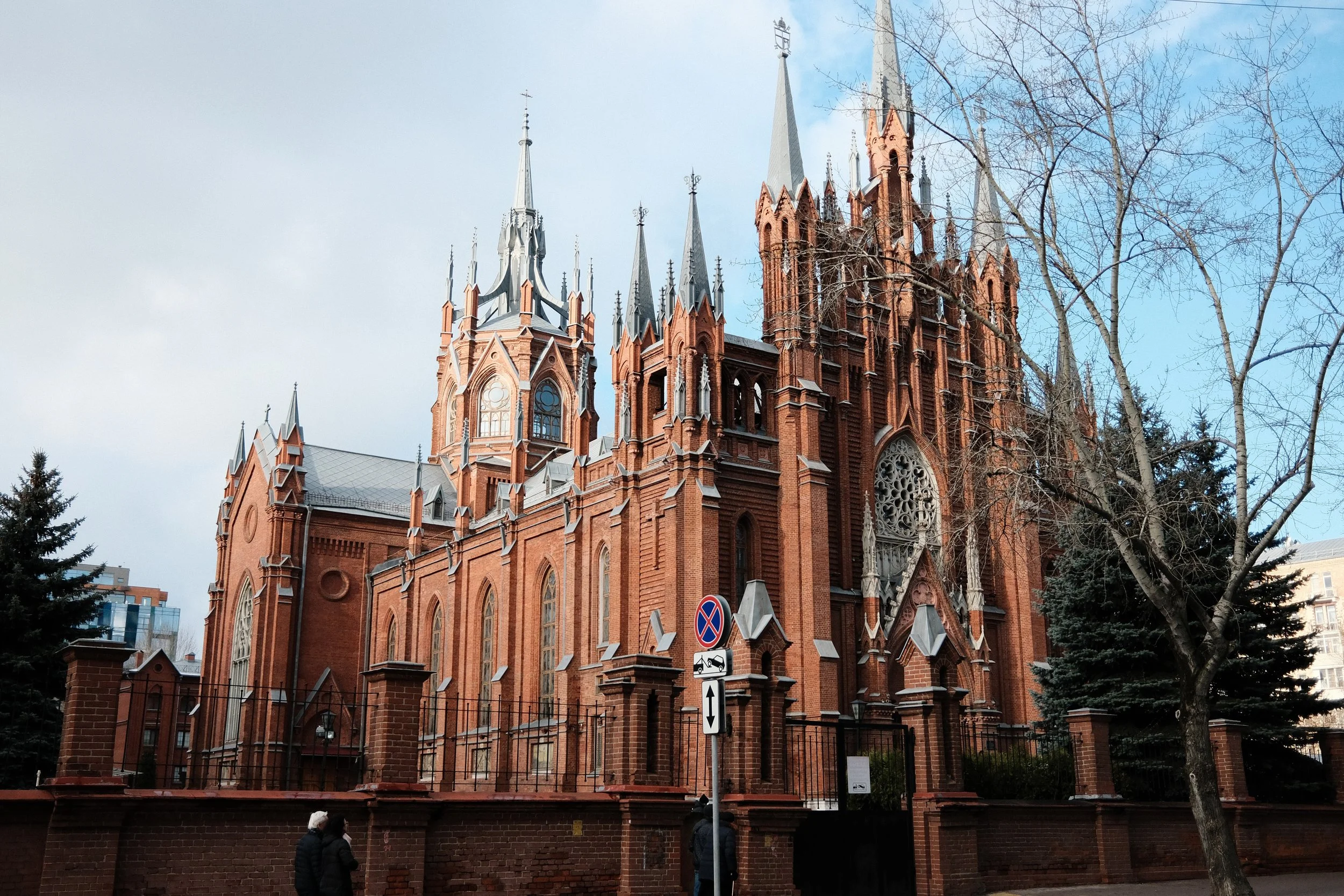 A large, red-brick Gothic-style church with multiple pointed spires and large stained glass windows, surrounded by trees and city buildings in the background.
