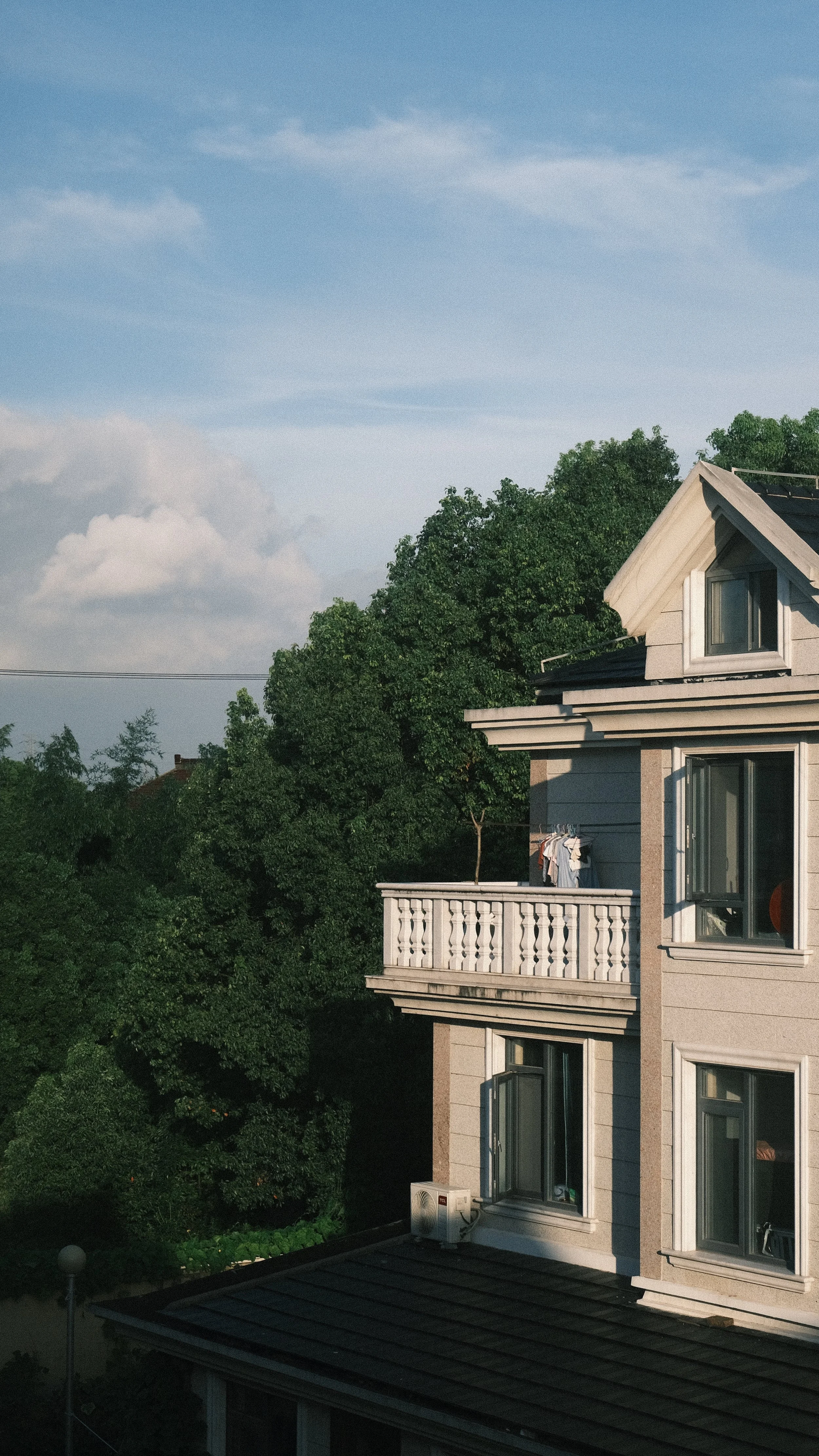 Residential building with windows and a small balcony, surrounded by lush green trees under a blue sky with clouds.