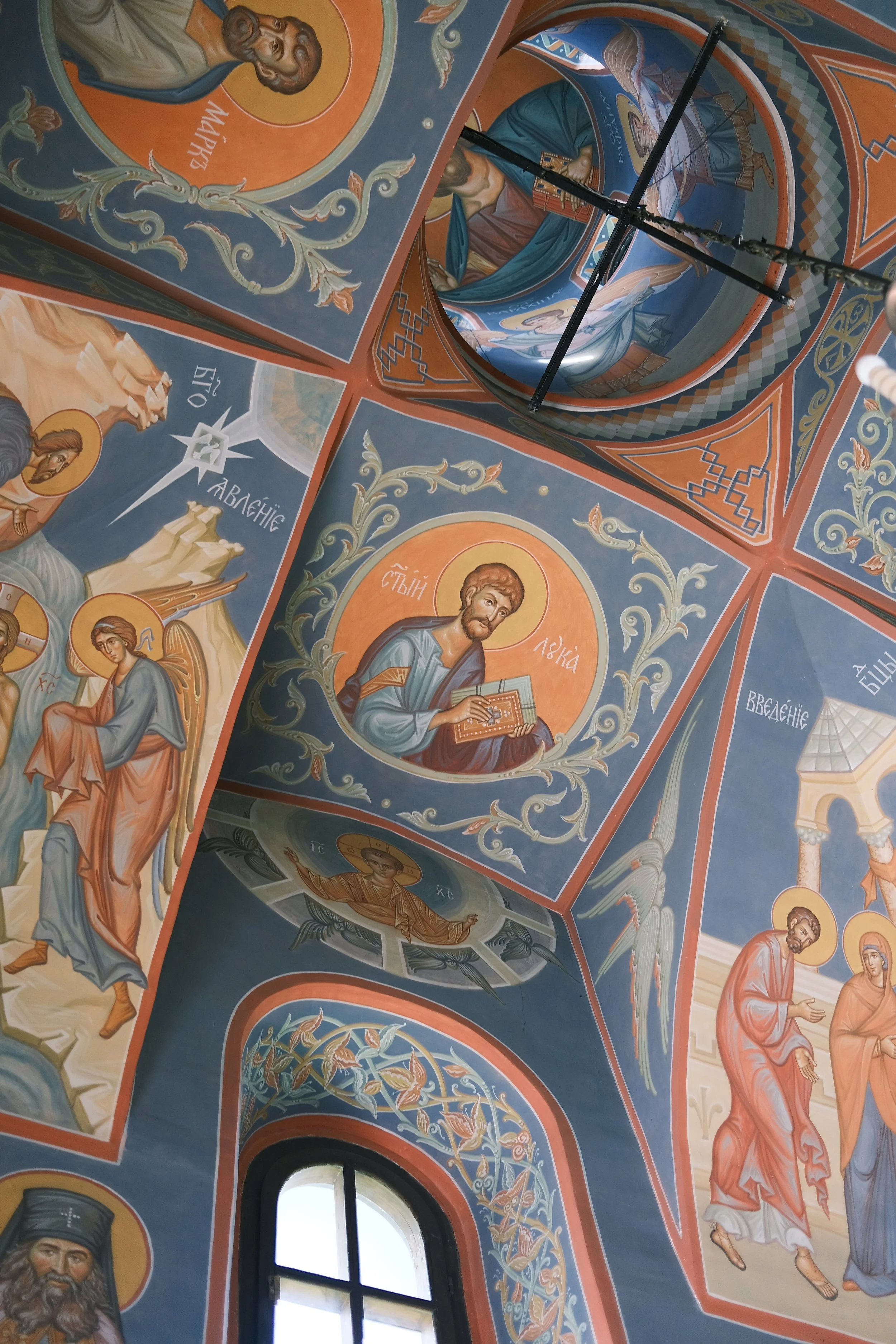 Colorful religious frescoes painted on the ceiling inside an Orthodox Christian church, including images of saints and biblical scenes with Greek inscriptions.