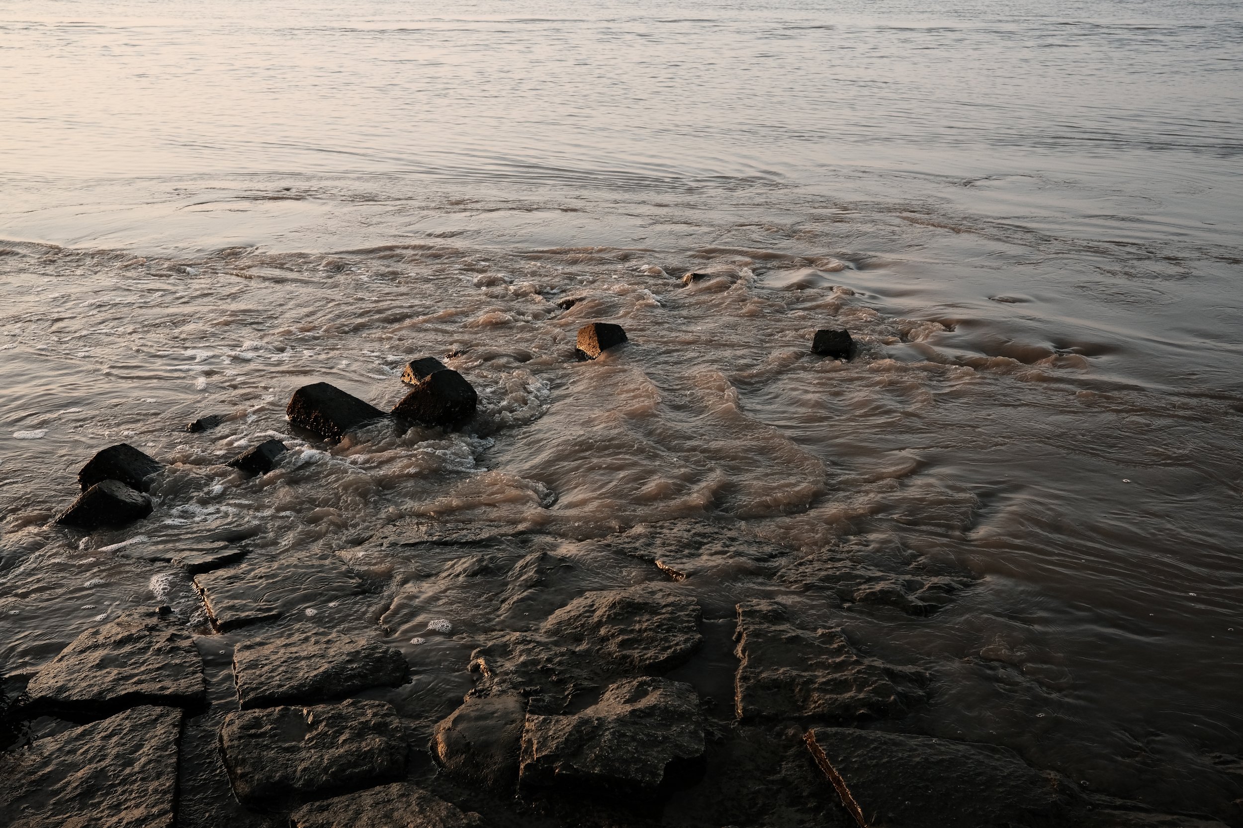 Rocks and concrete blocks along a shoreline with water gently lapping at the edges during sunset.